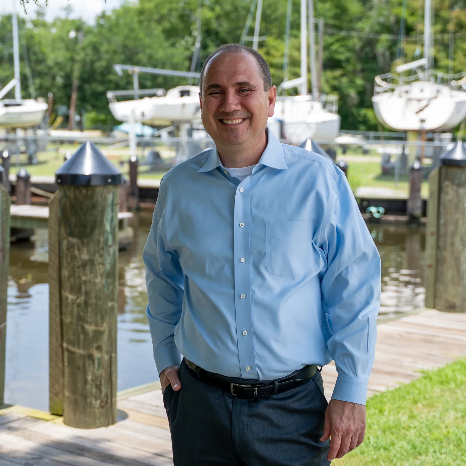 Smiling man in blue shirt standing on wooden dock by marina with boats and trees in background