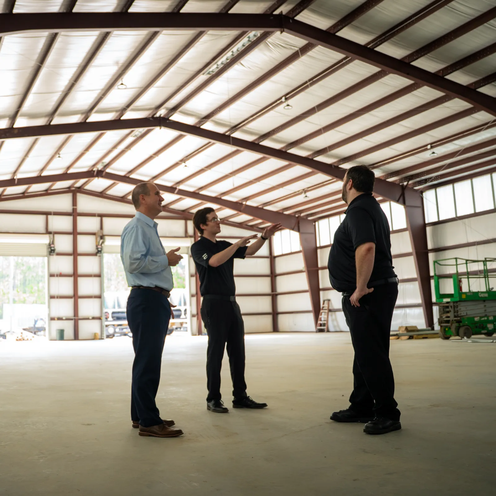 Three men discussing plans inside a large open industrial warehouse with metal beams and concrete flooring.