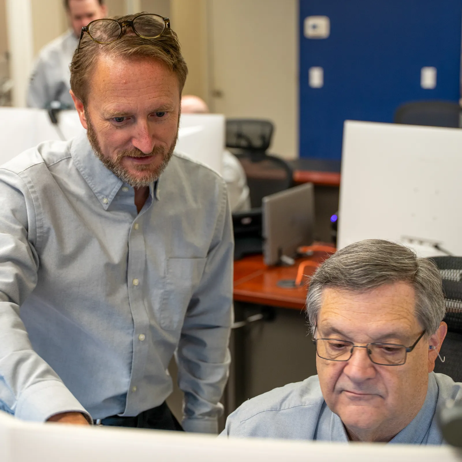 Two men collaborate at a computer workstation in a modern office environment with technology and blue walls.