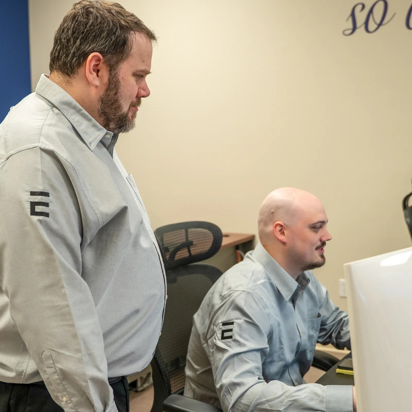 Two men wearing matching gray shirts working together on a computer in an office setting.