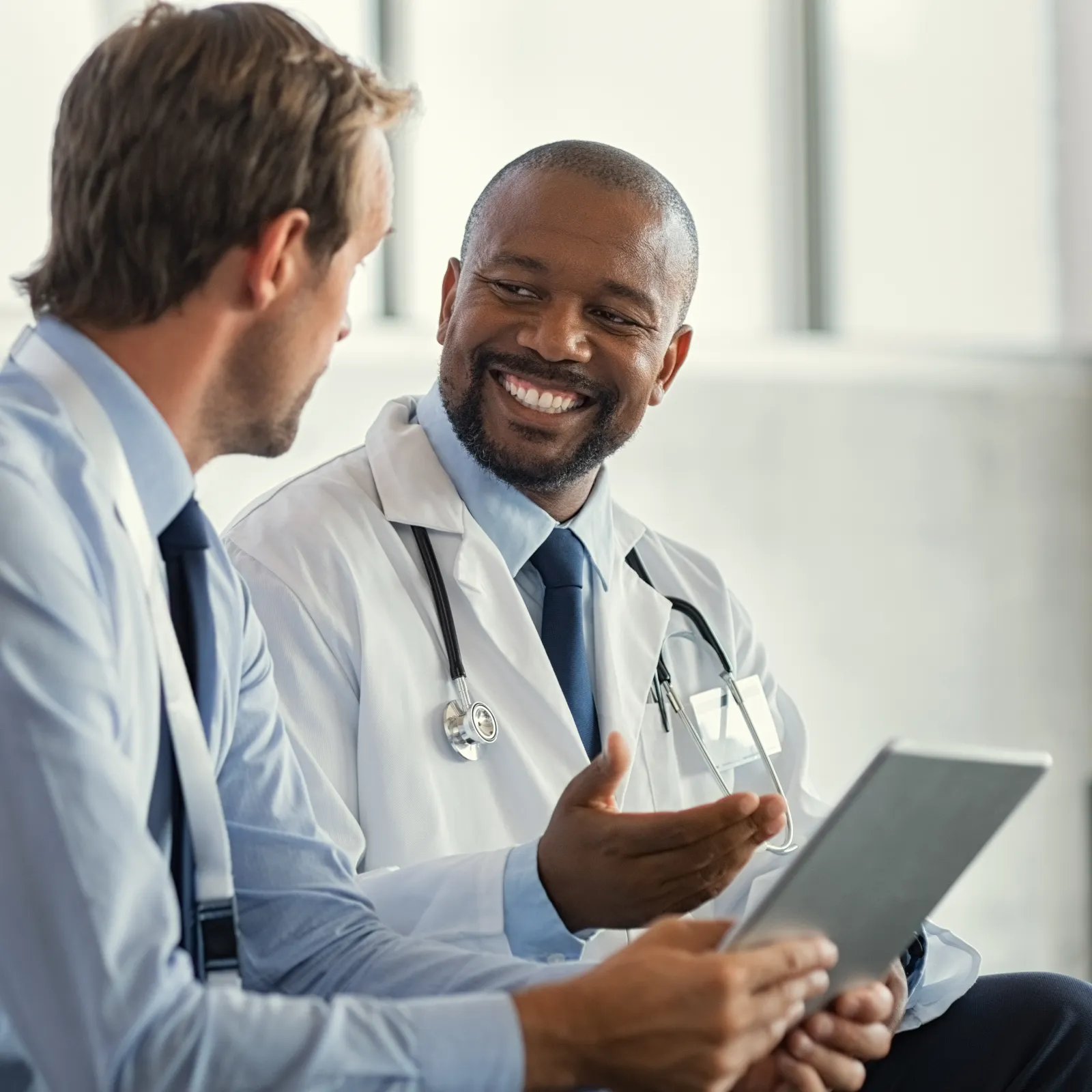Doctor in white coat with stethoscope discussing health information with patient using tablet device.