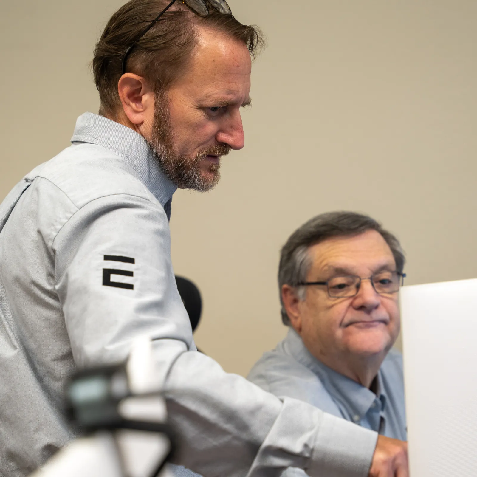 Two men collaborating at a computer, one pointing at the screen while the other looks on attentively in an office setting.