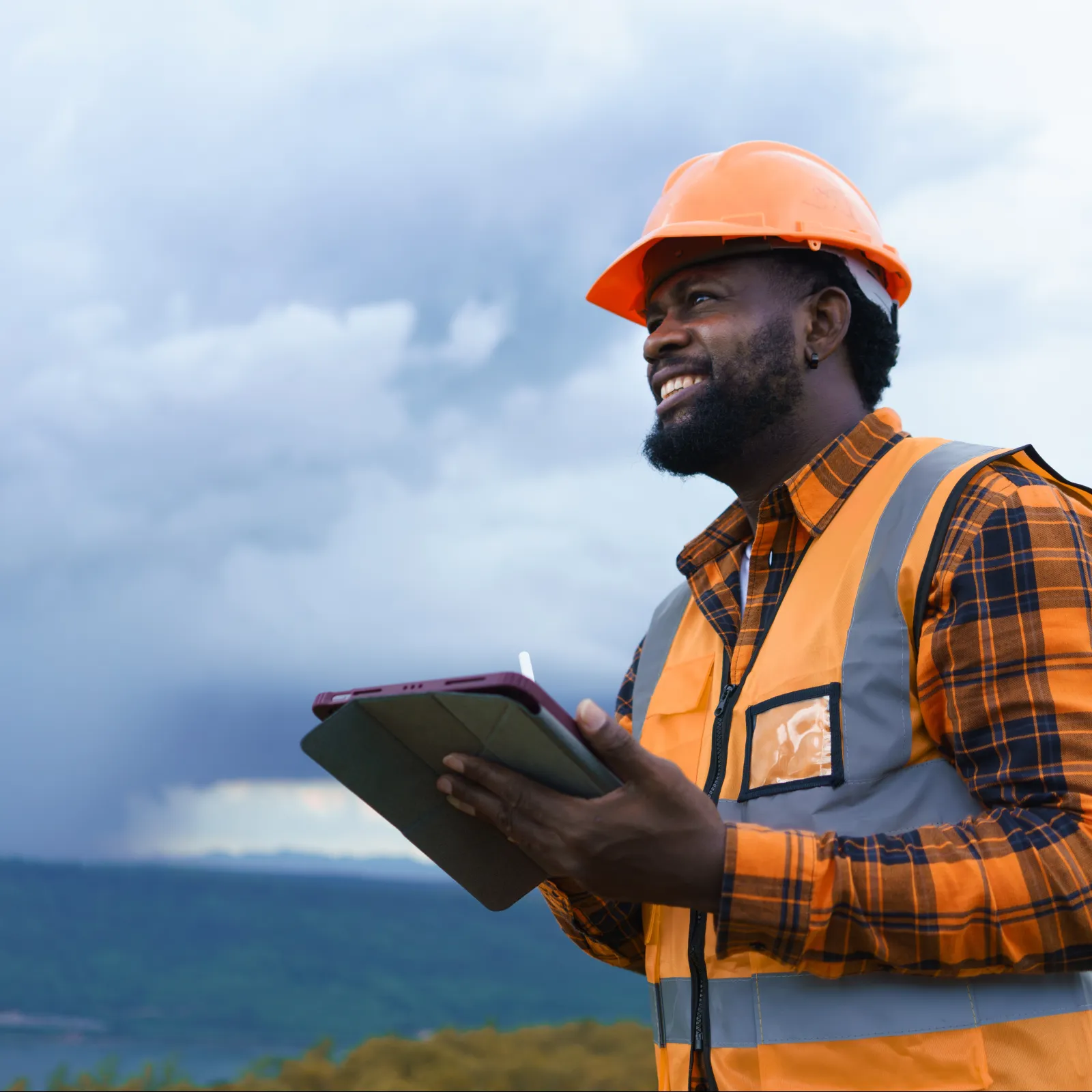 Construction worker in orange safety gear holding tablet, smiling outdoors with cloudy sky and hills in background