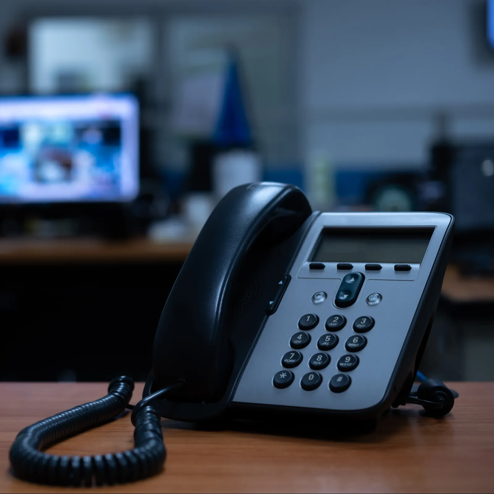 Desk phone with corded handset in focus on office desk with blurred computer monitors in background