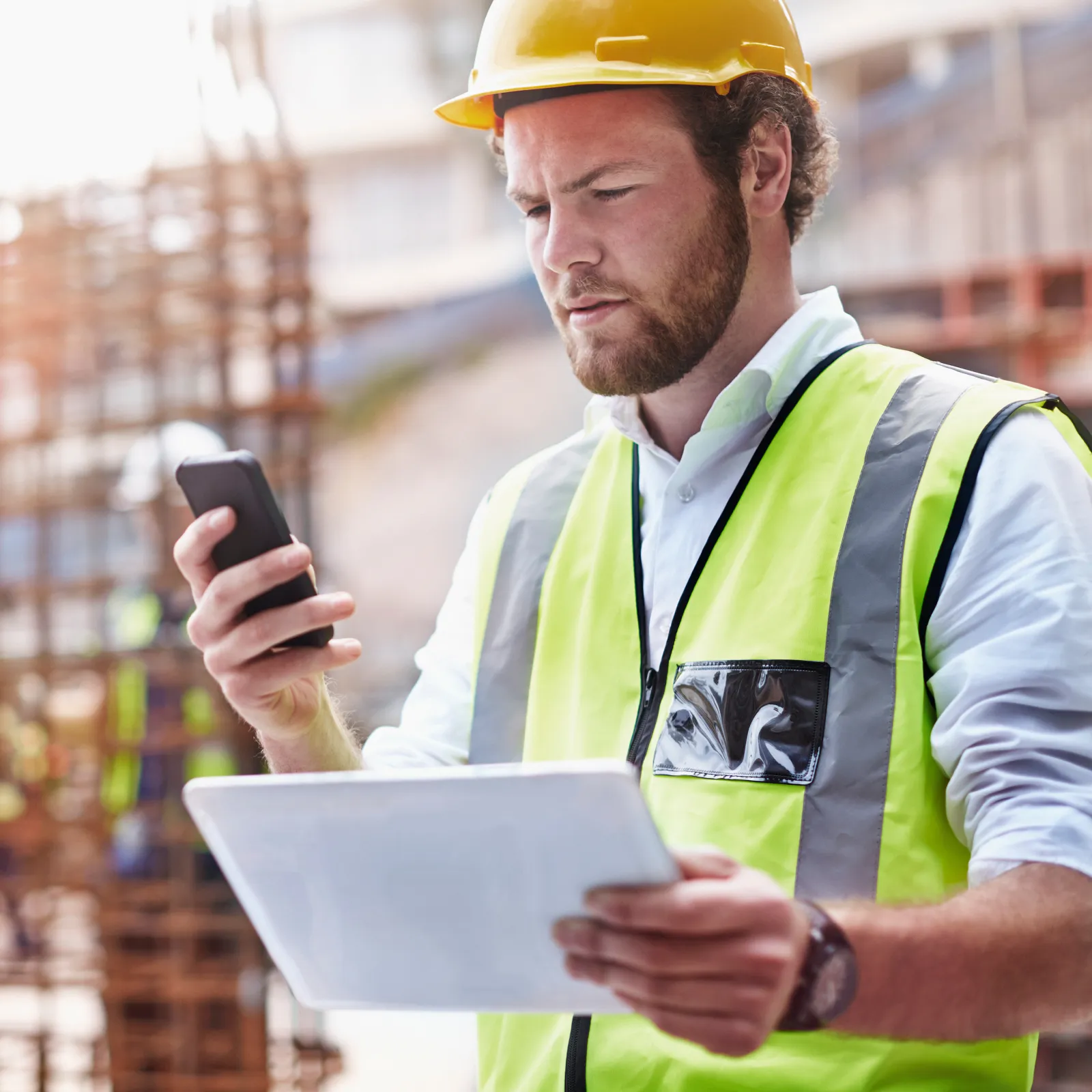 Construction worker in safety gear checks smartphone and tablet at building site with steel framework in background