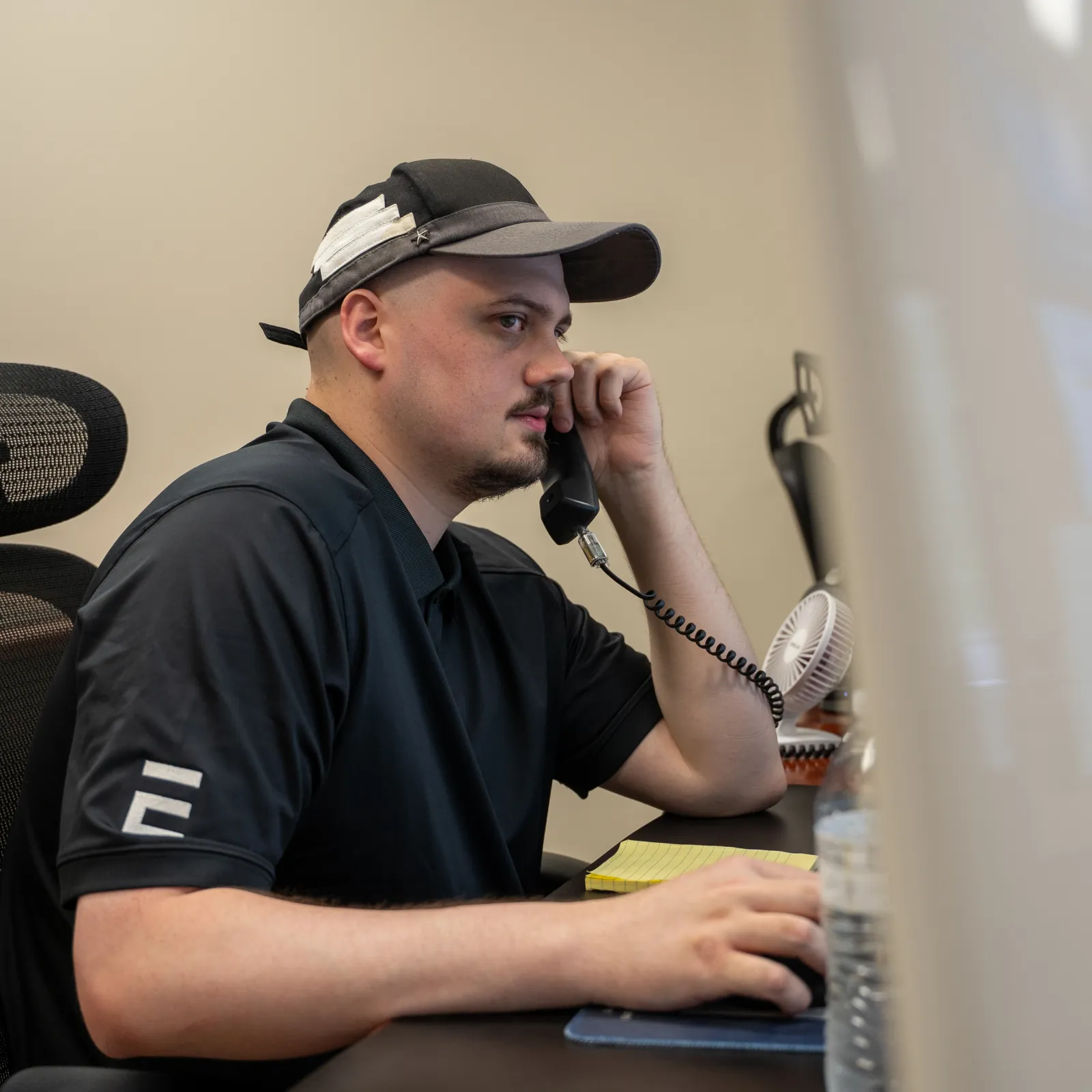 Man in black shirt and cap talks on office phone while working at computer desk with large monitor.