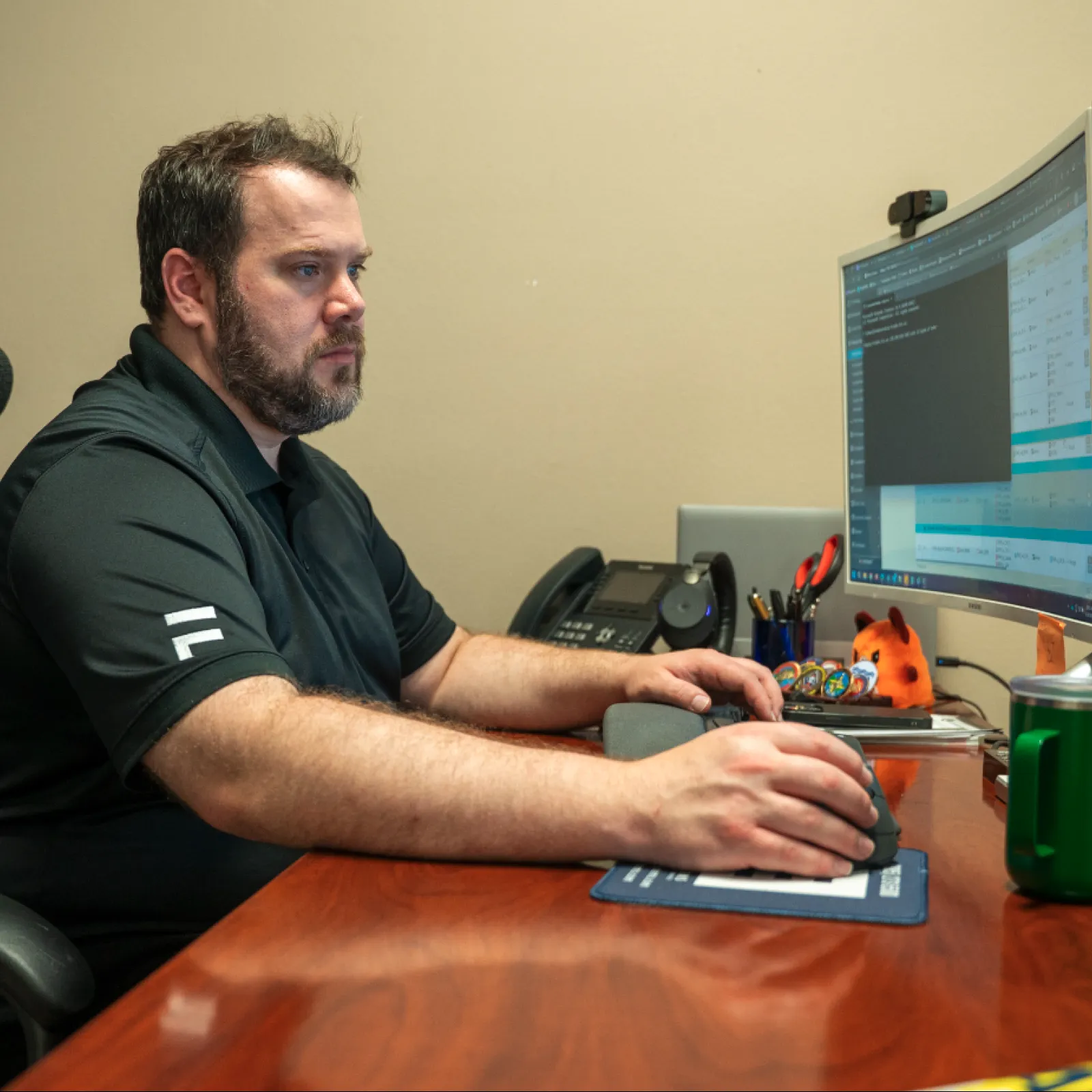 Man in black shirt working on a dual-monitor computer setup in an office with wooden desk and green mug.