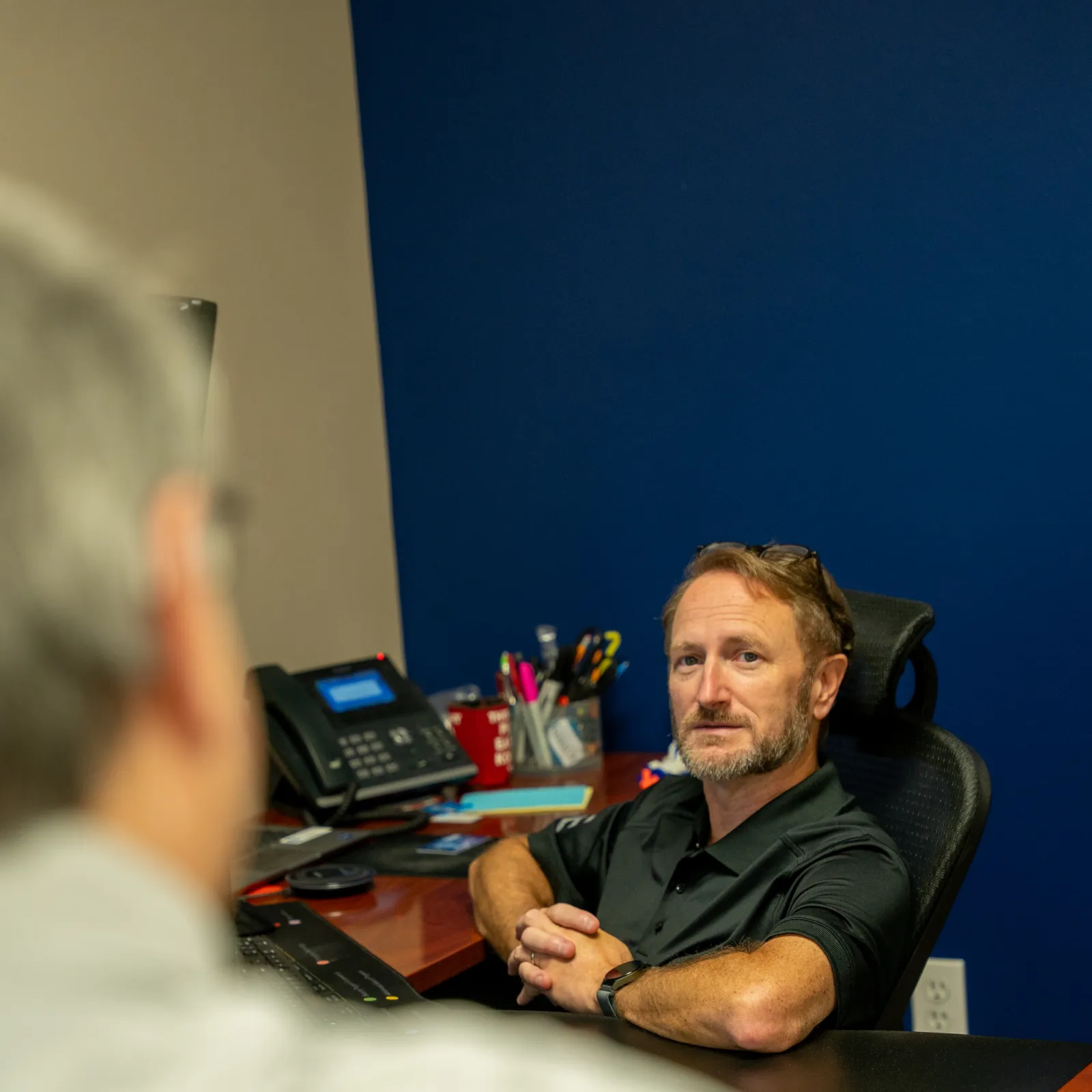 Man with beard sitting at desk in office, engaged in conversation with blurred person in foreground.