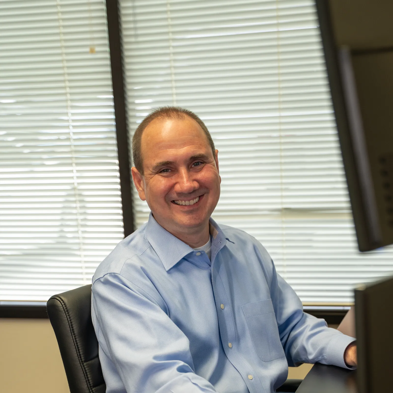 Smiling man in blue shirt working on computer at office desk with window blinds in background