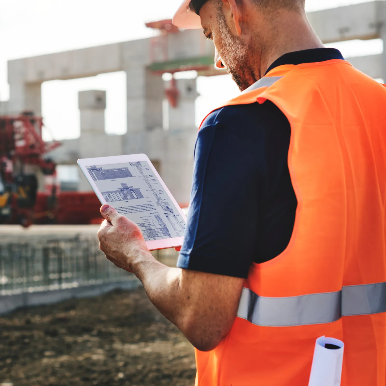 Construction worker in orange safety vest reviewing digital blueprints on a tablet at a busy building site.