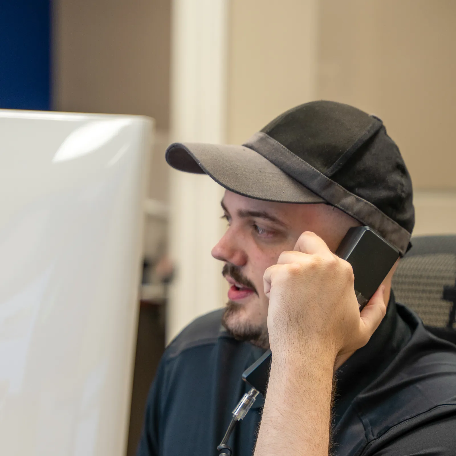 Man in black cap and shirt talking on a desk phone while working at a computer in an office.