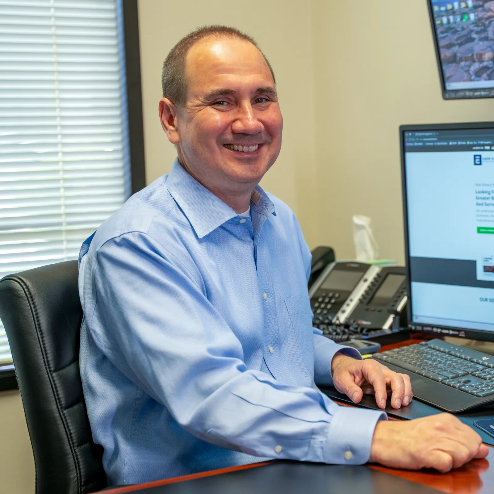 Smiling man in blue shirt sitting at desk with computer monitors and office phone in bright modern office.