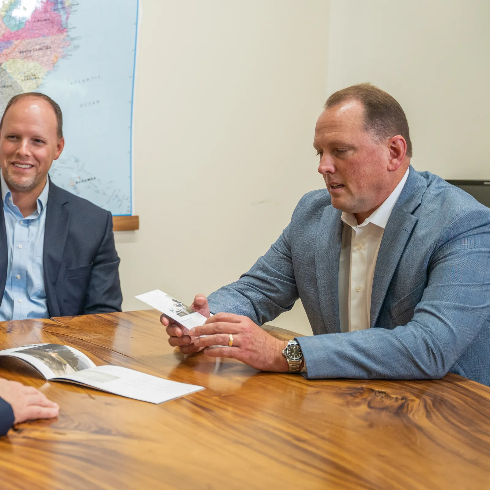Three businessmen in suits discussing documents at a wooden table with a US map on the wall.