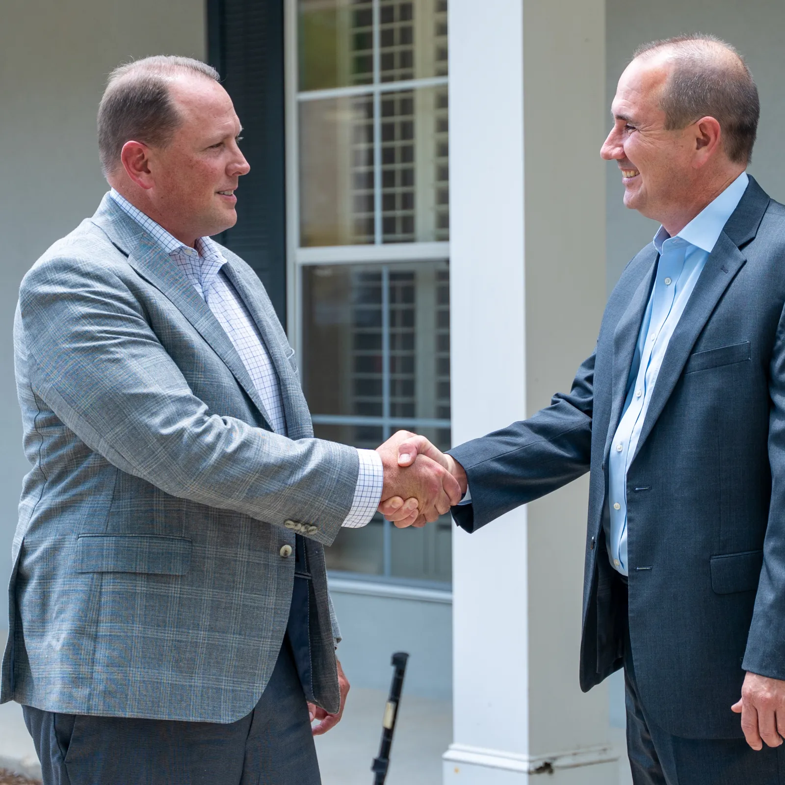 Two professional men shaking hands outside a building, dressed in business attire with smiles, indicating agreement.