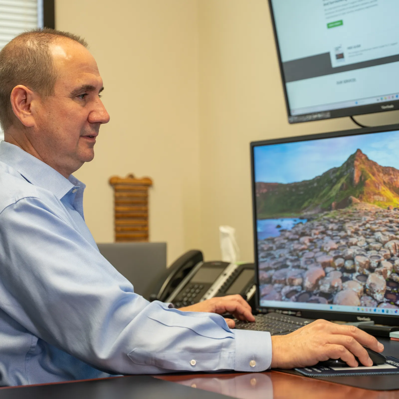 Man in blue shirt working at a desk with multiple monitors, one displaying a rocky coastal landscape.