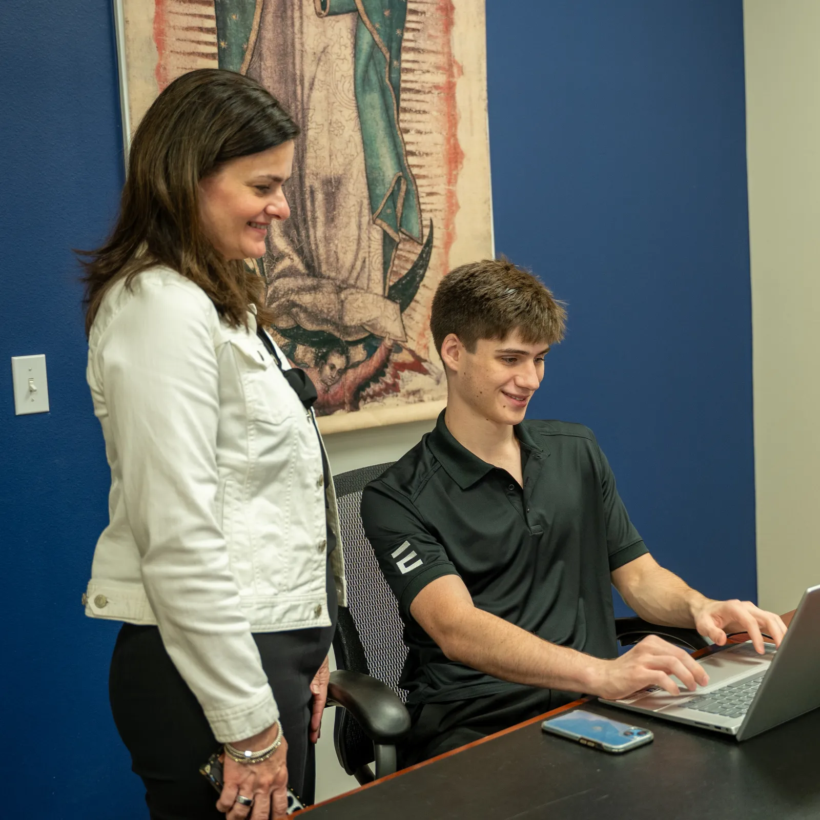 Young man typing on laptop while woman stands next to him smiling in office with blue wall and artwork.