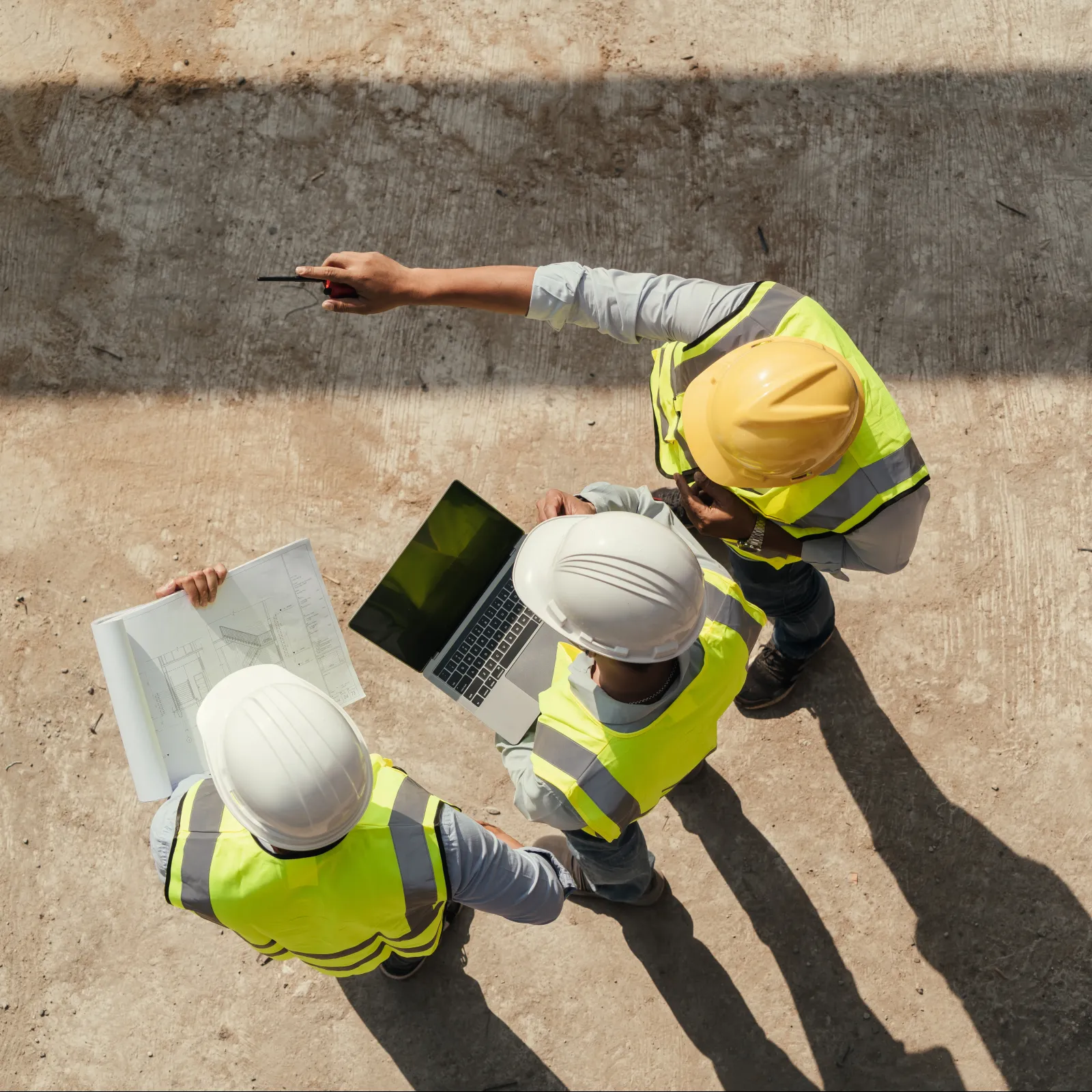 Top view of three construction workers with helmets and vests reviewing plans and laptop on site.