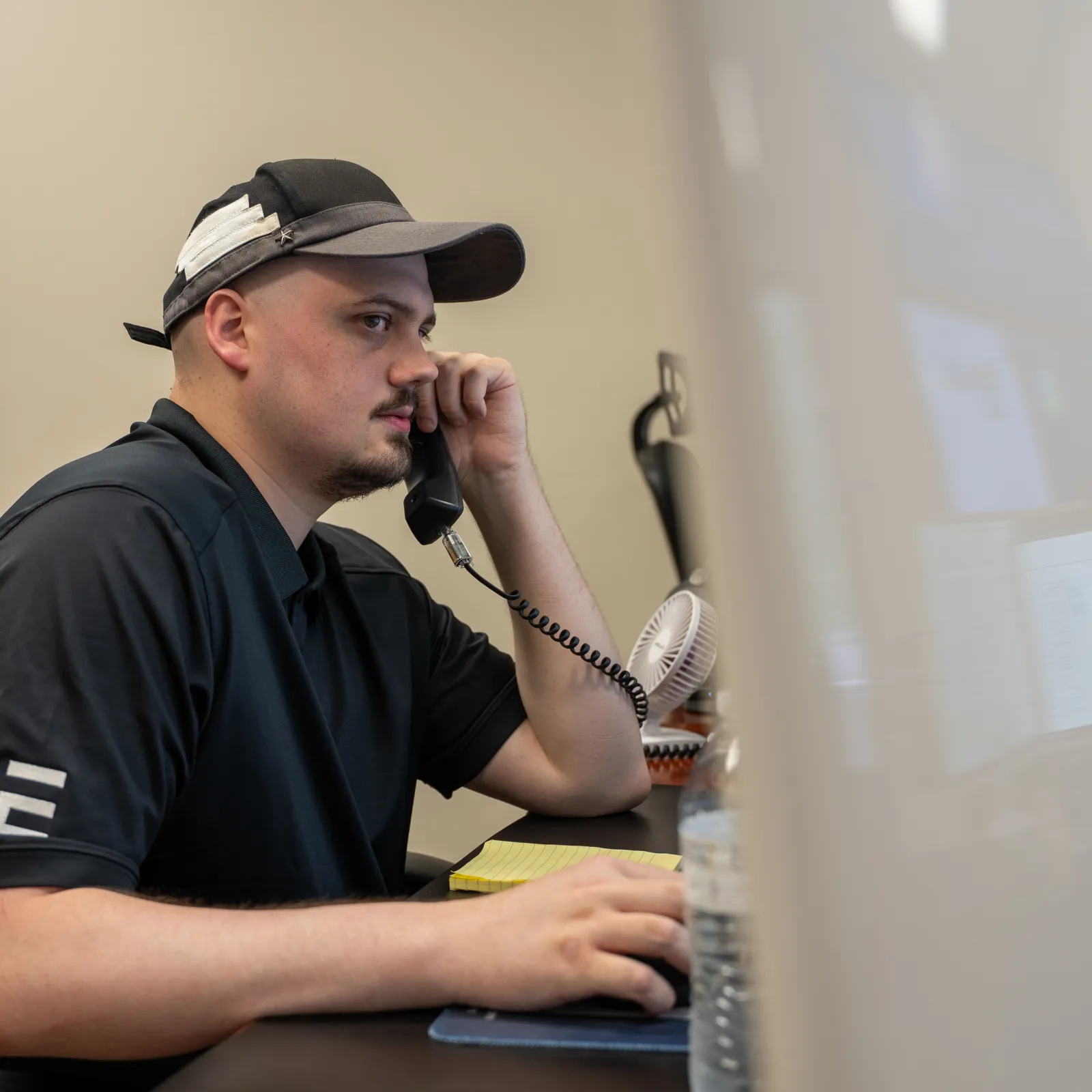 Man in black cap and shirt speaking on phone while working at a computer in an office.