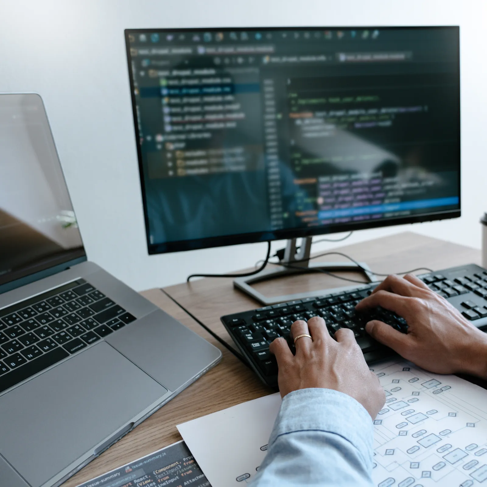 Person typing code on a keyboard with laptop and monitor displaying software code on a wooden desk.