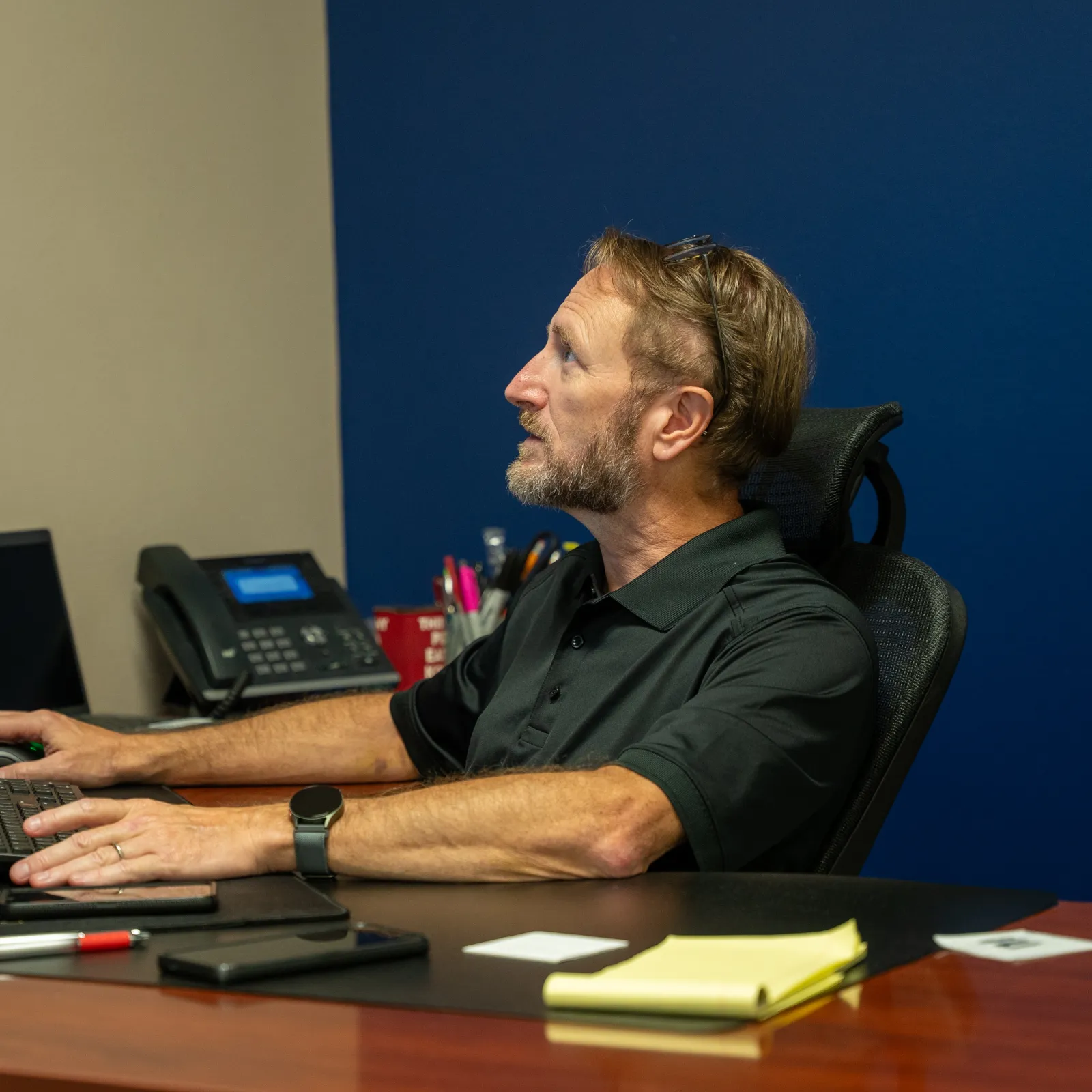 Man in black shirt working at a desk with multiple monitors, phone, and office supplies in a modern workspace.