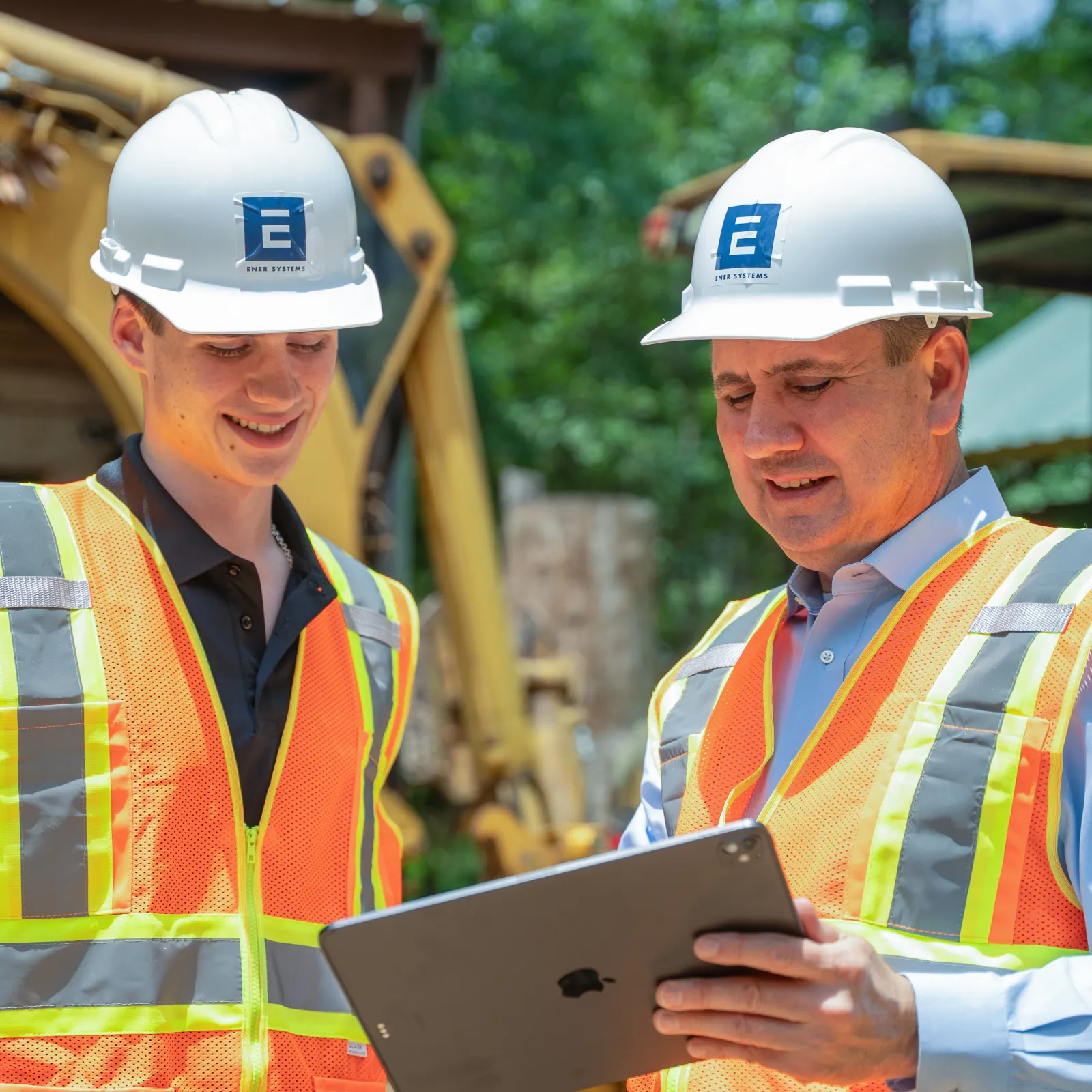 Two construction workers wearing helmets and safety vests reviewing plans on a tablet at an outdoor site.