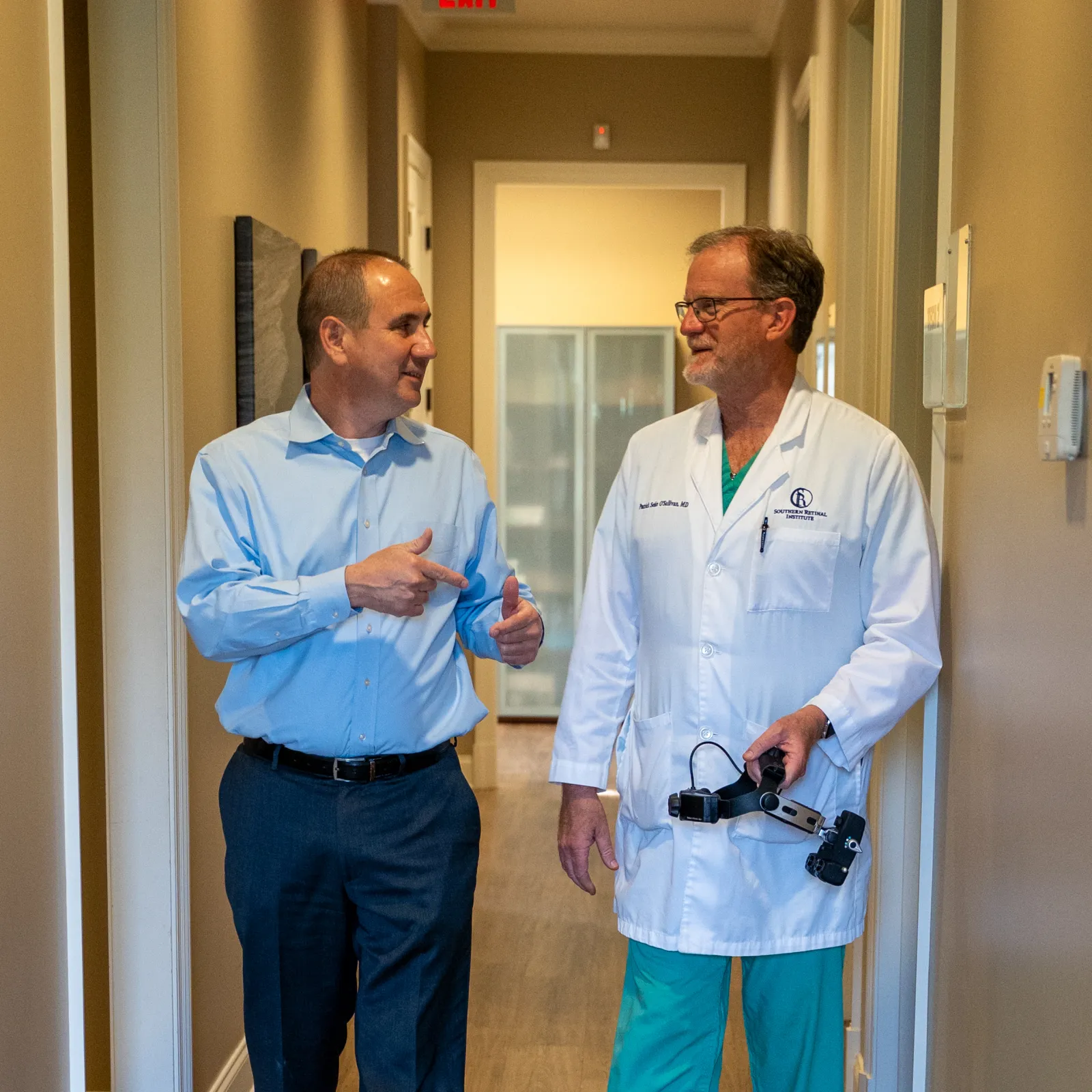 Two men walking and talking in a medical office hallway, one in business attire and one doctor in a white coat