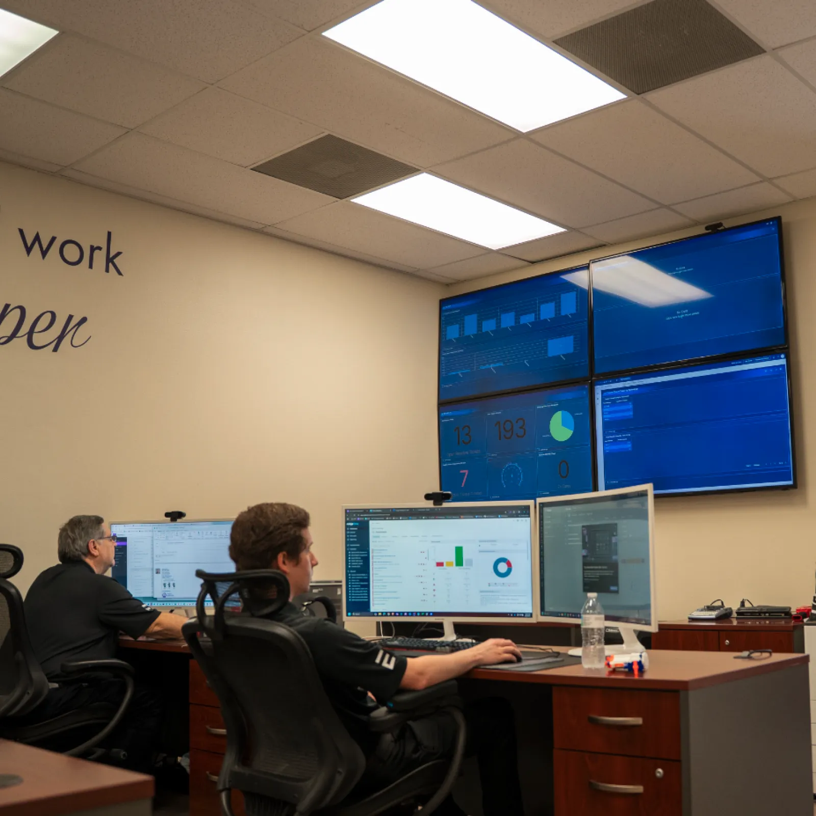 Two men monitoring multiple computer screens and wall-mounted displays in a modern office workspace.