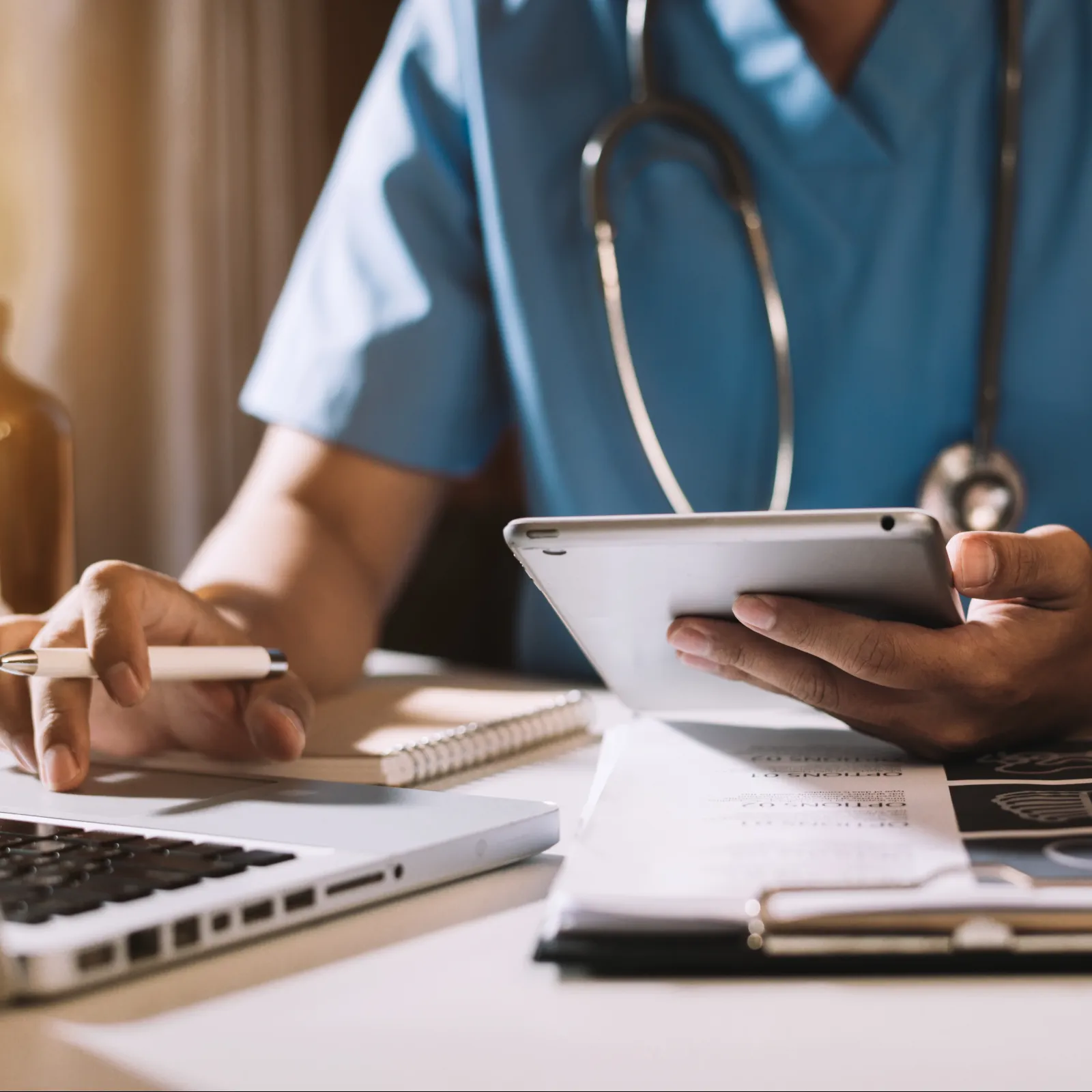 Doctor in blue scrubs using tablet and laptop with medical documents on desk in warm light.