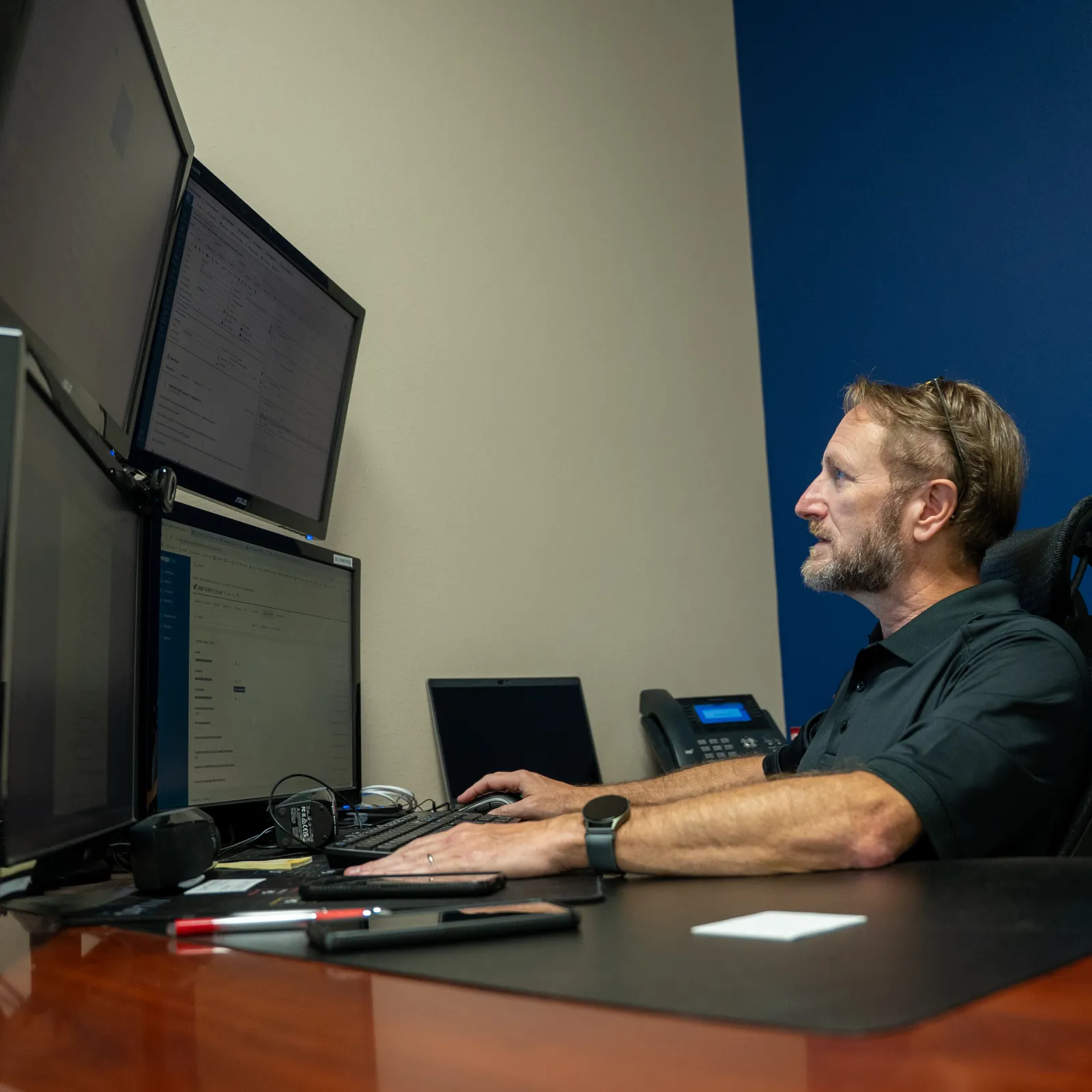 Man with beard working on multiple computer monitors at desk in modern office with blue and white walls