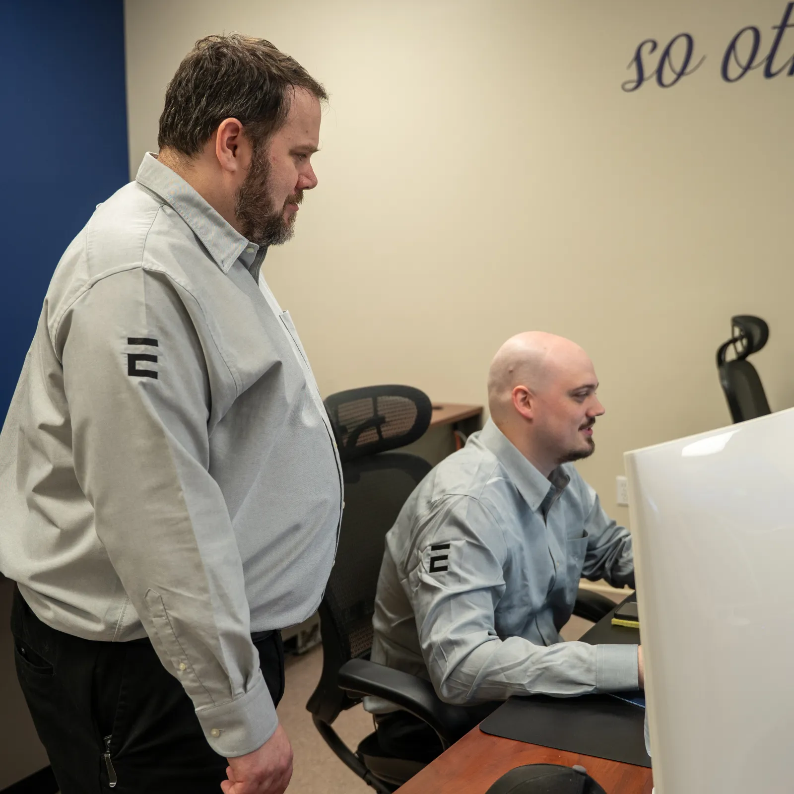 Two men in matching uniforms working together at computer workstations in an office with religious artwork on the wall.