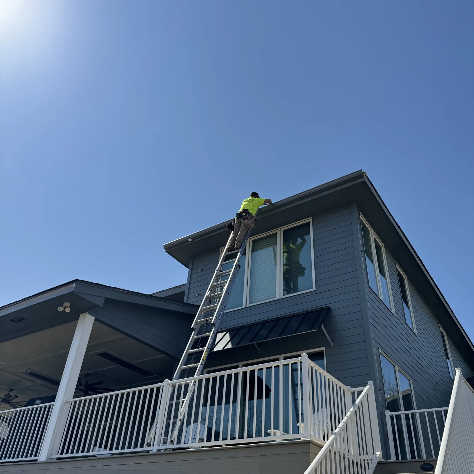 Worker in a neon shirt on a ladder repairing or inspecting the roof of a gray two-story house on a clear sunny day