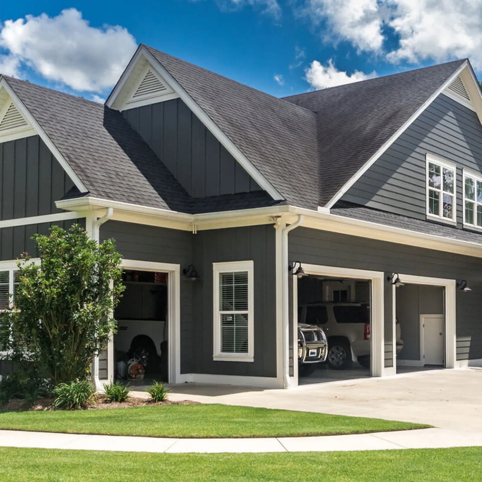 Modern two-story house with dark gray siding, white trim, multiple garages, and a neatly manicured lawn.