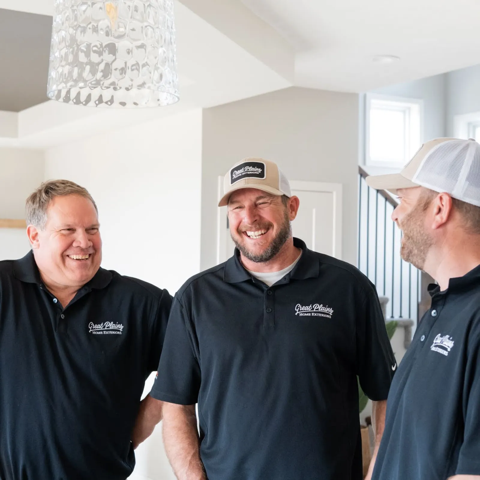 Three men wearing Great Plains Home Exteriors black polos and hats smiling and discussing color samples indoors.