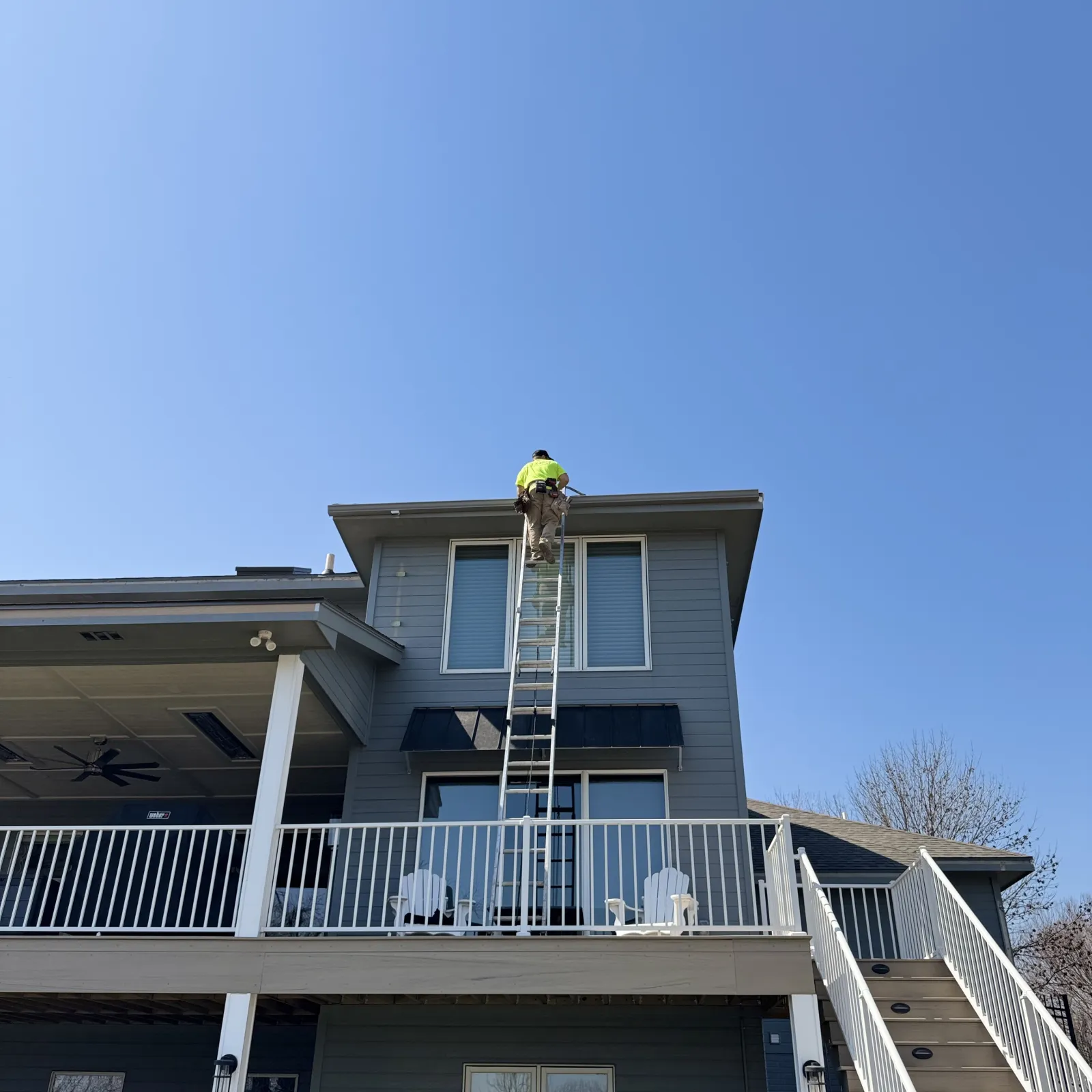 Worker in a neon jacket climbs ladder to roof of two-story gray house on a sunny day.