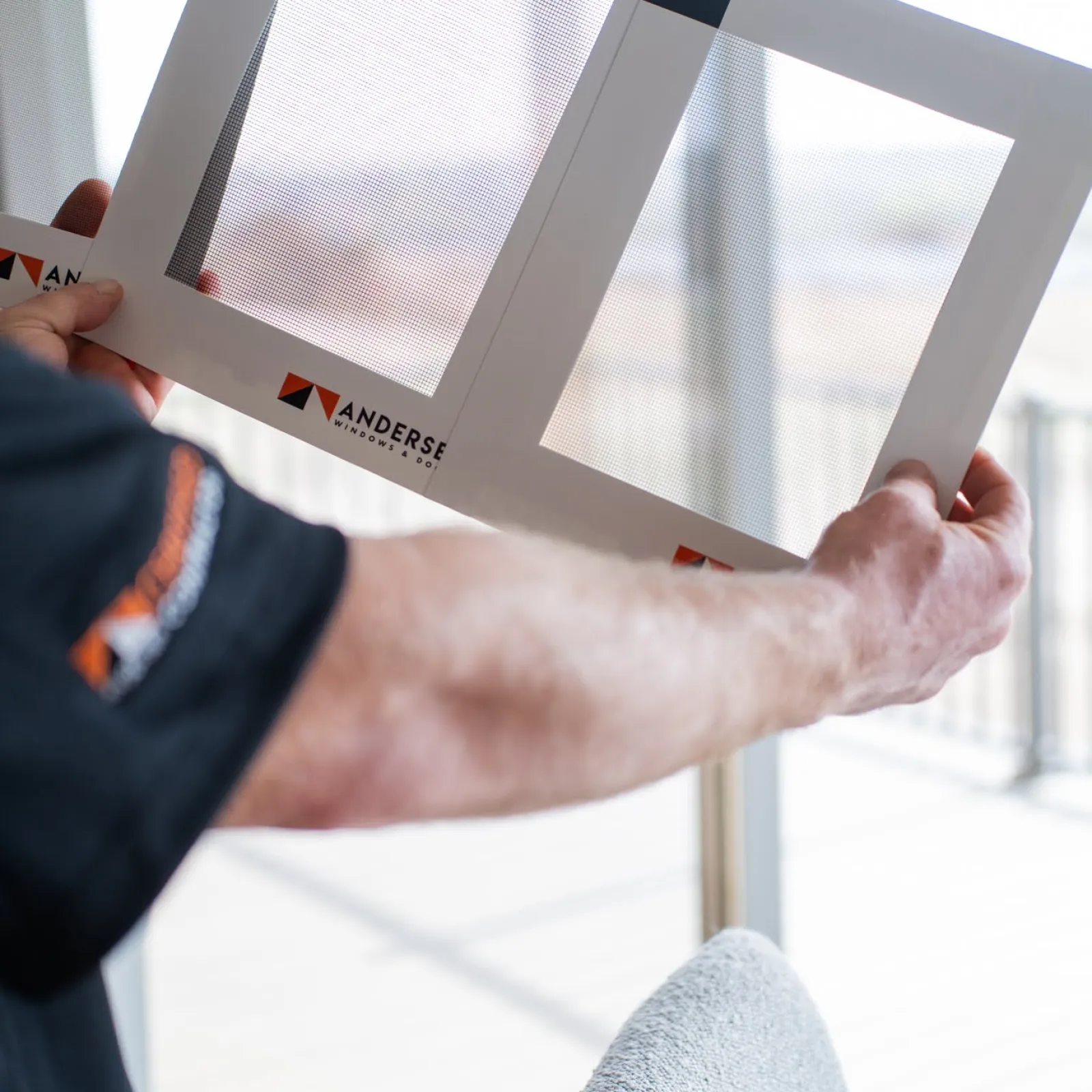 Person holding window screen samples labeled fiberglass and aluminum by Anderse Windows and Doors