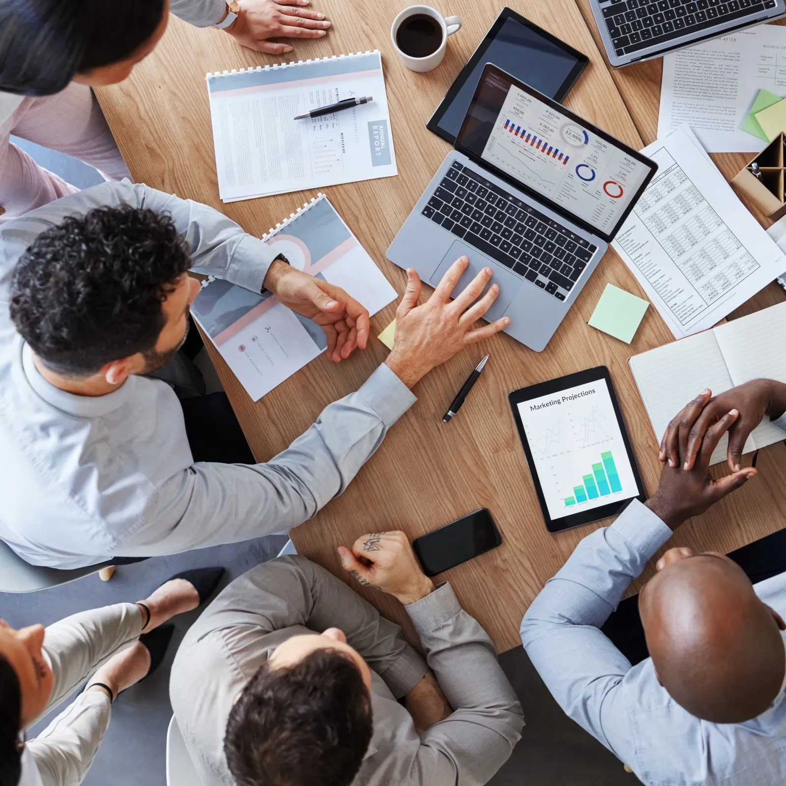 Top view of diverse team discussing marketing data with laptops, tablets, charts, and notes during meeting.