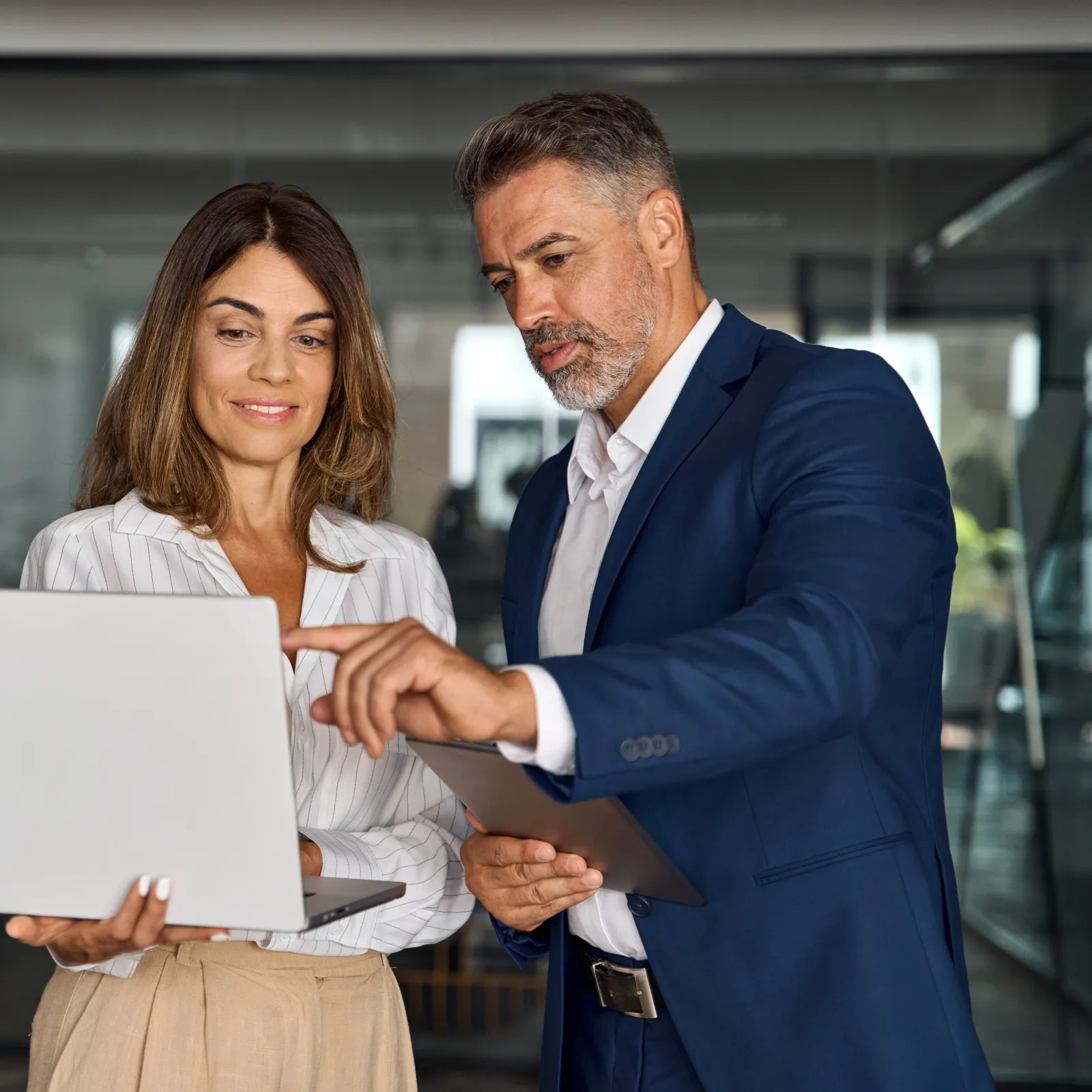 Professional man and woman discussing work while looking at a laptop in modern office hallway.