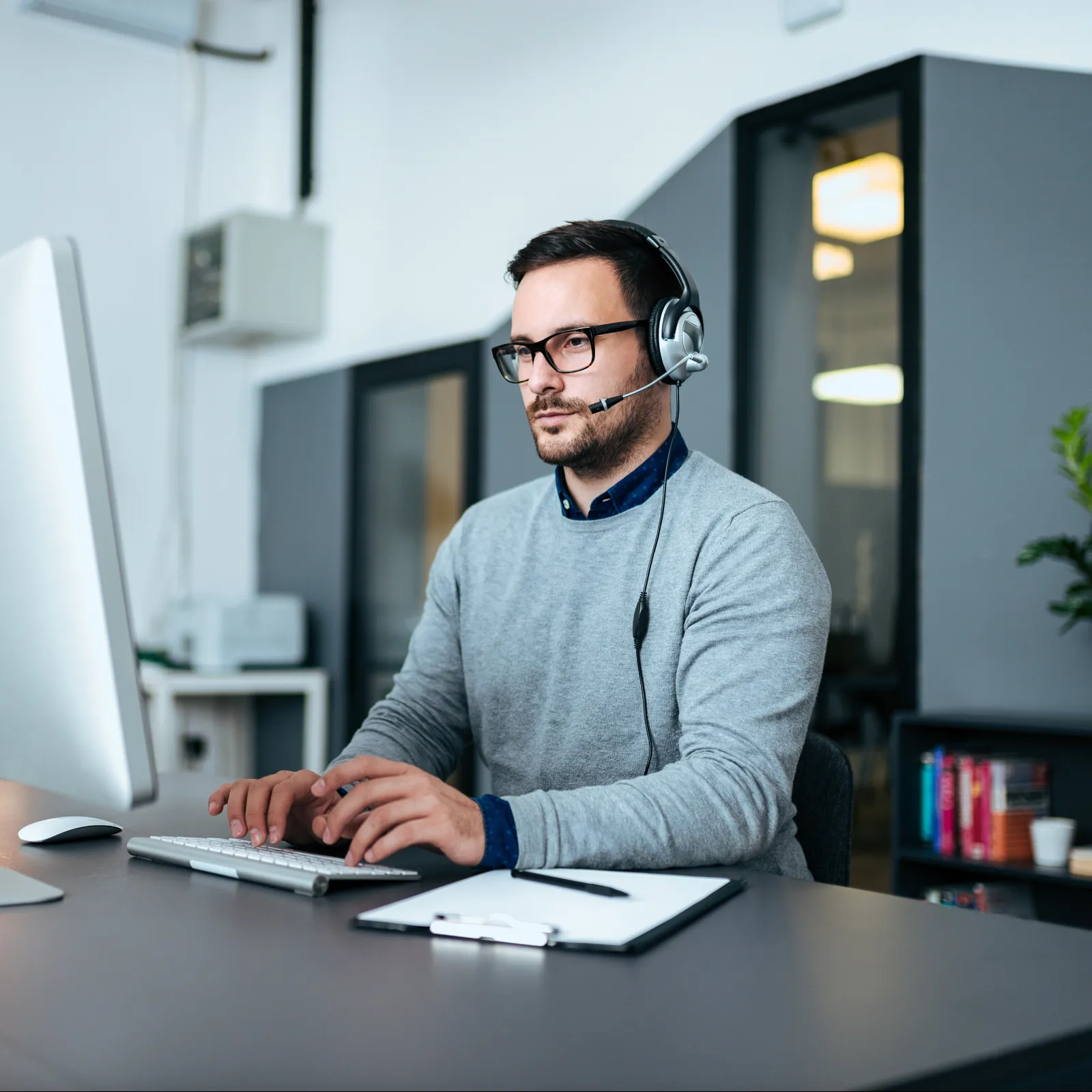 A focused man with headphones works at a desktop computer in a modern office setting.