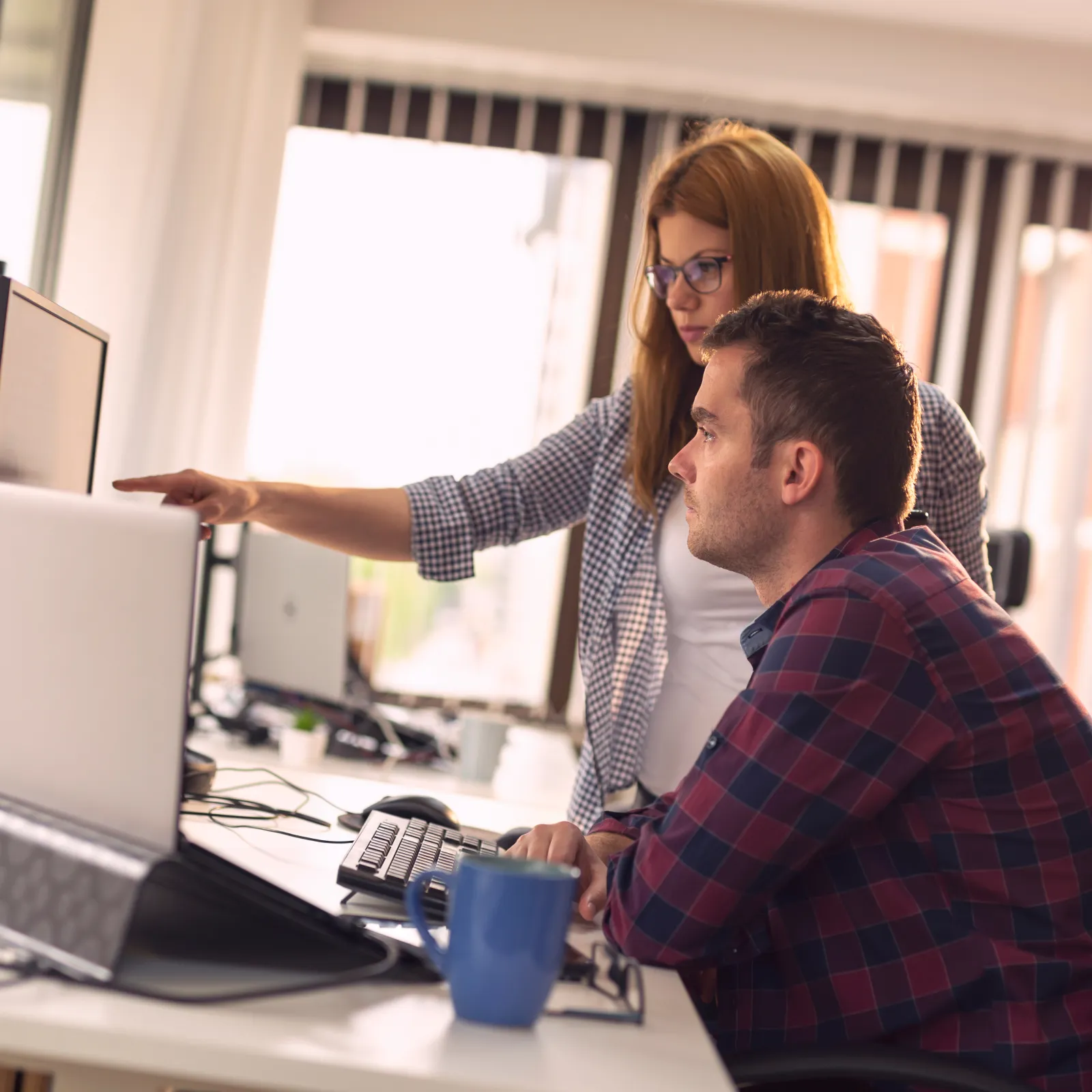 Two colleagues collaborating at a desk with computer screens in a modern office space.