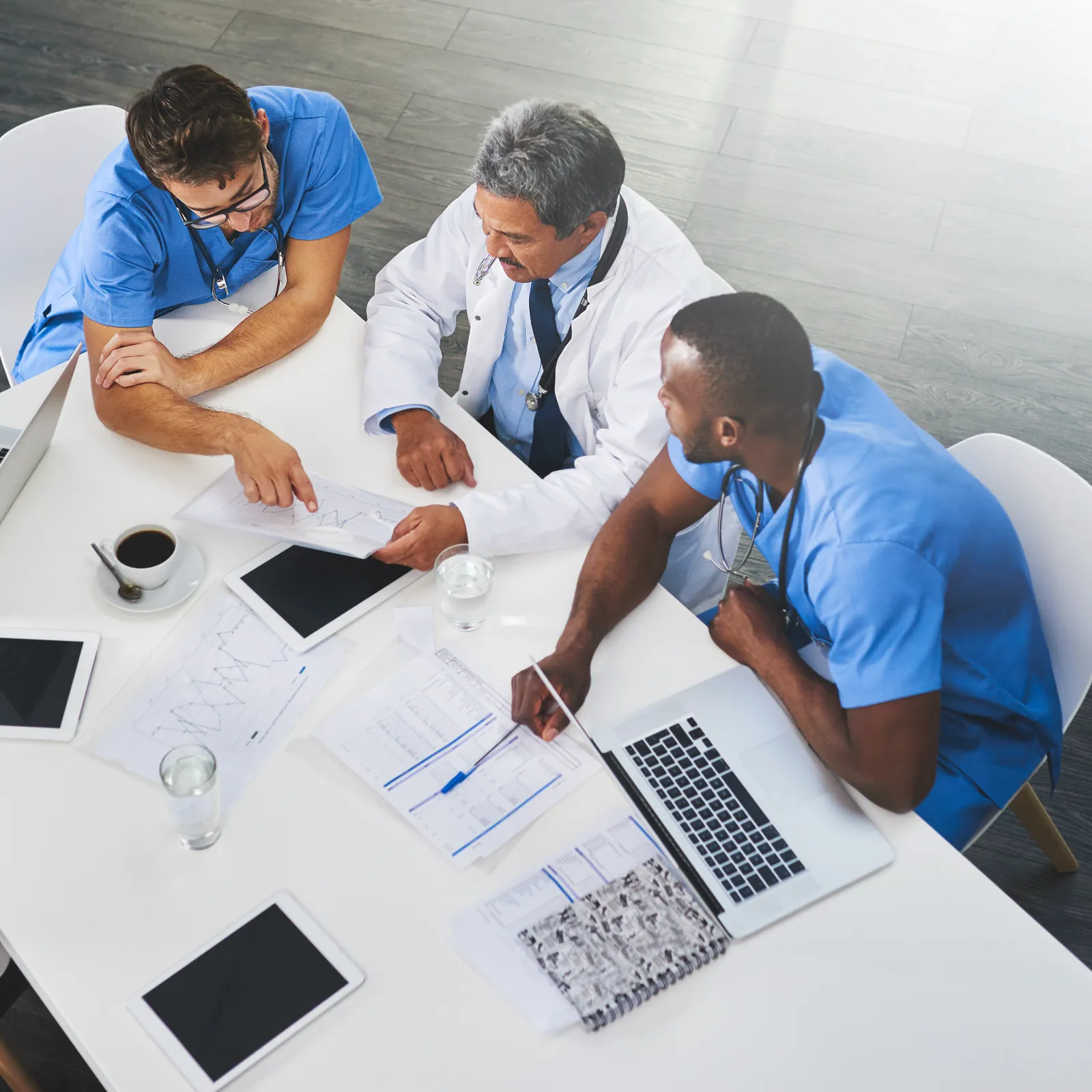 Medical team collaborating around a table with laptops, charts, and coffee in a bright, modern office.