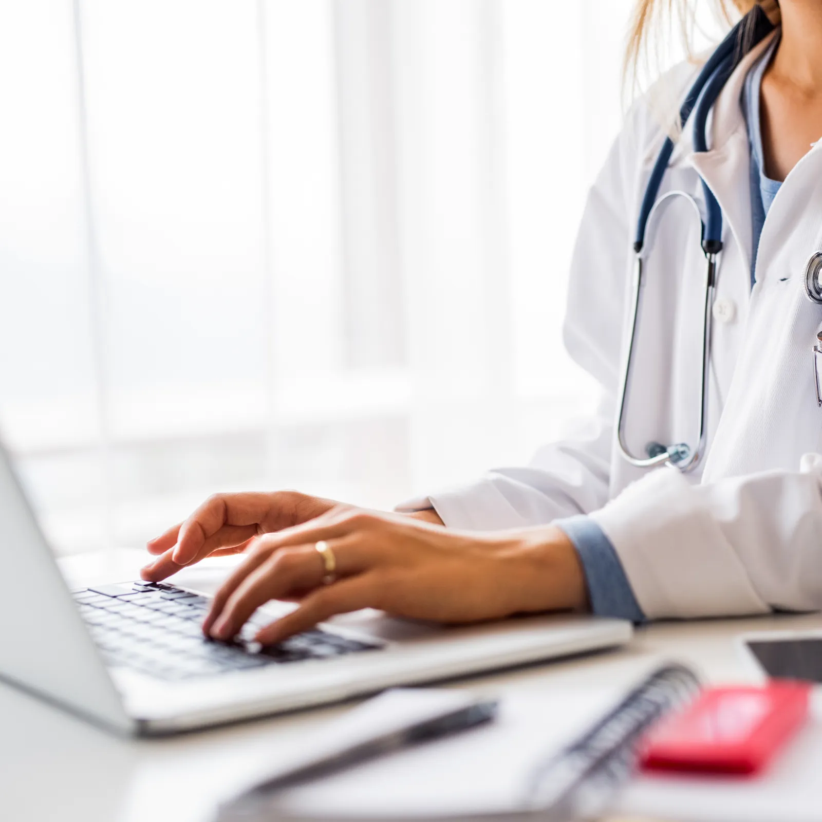 Healthcare professional in a white coat typing on a laptop at a desk.