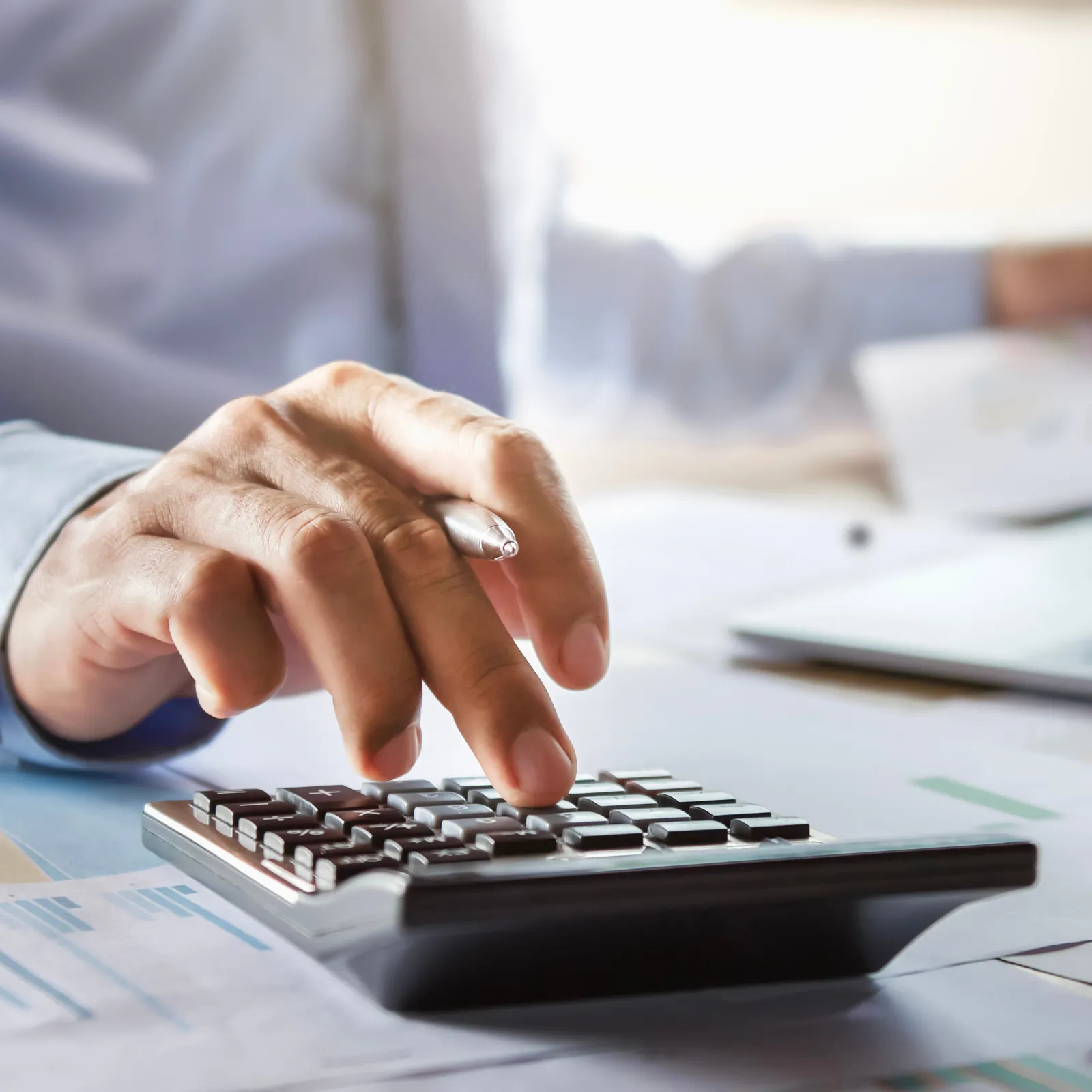 A person using a calculator while analyzing financial documents and a laptop on a desk.