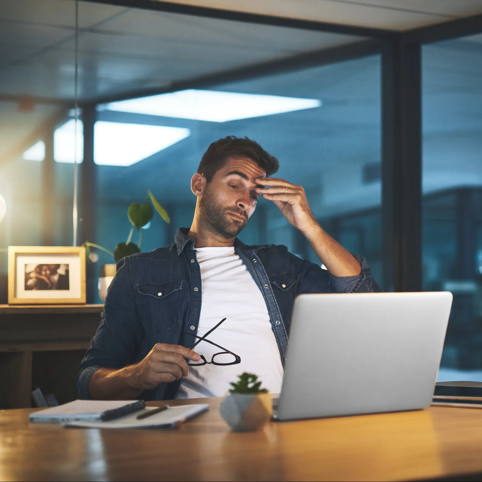 A man sitting at a desk in an office, stressed while working on a laptop at night.