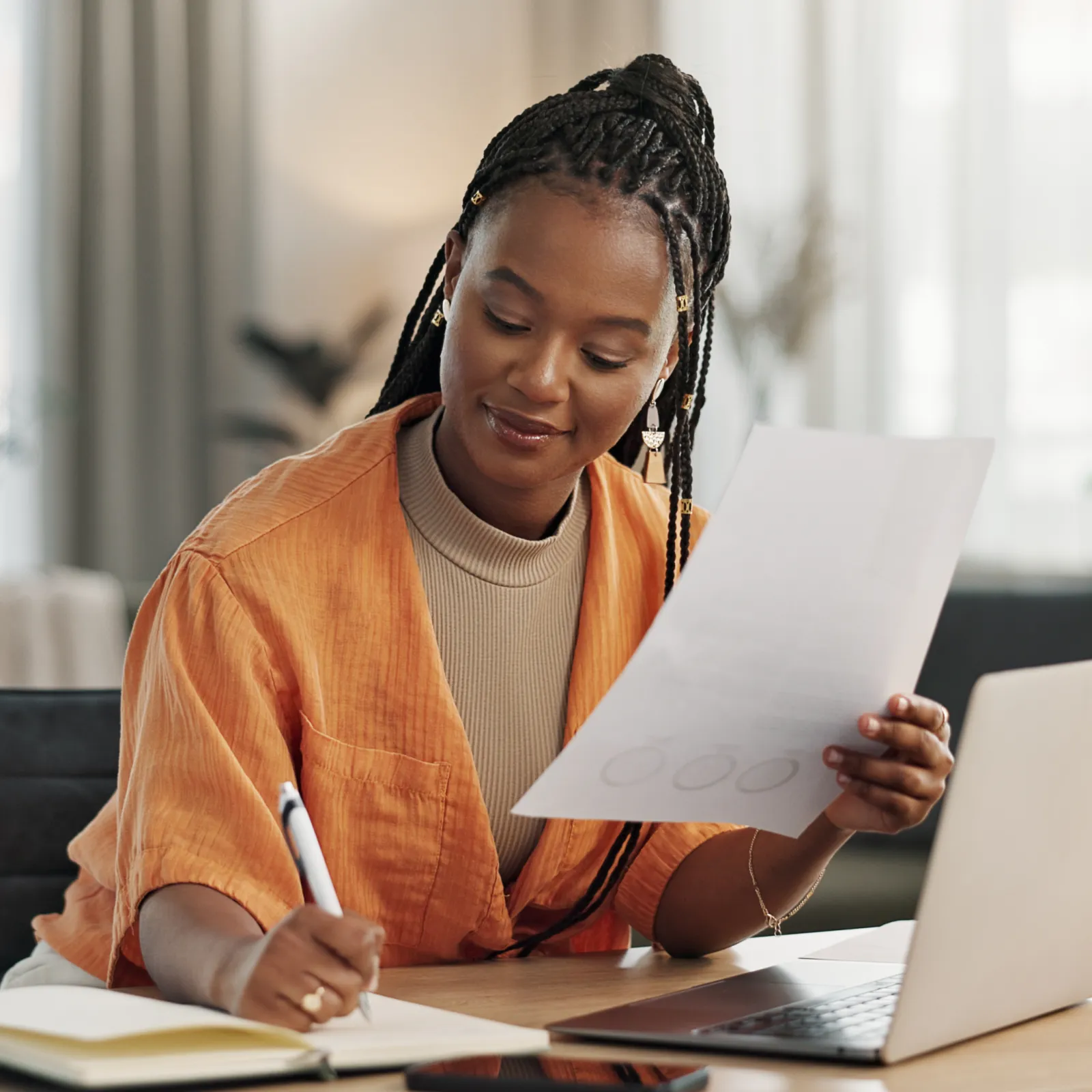 Woman in an orange cardigan takes notes at a desk with a laptop and papers in a bright office.