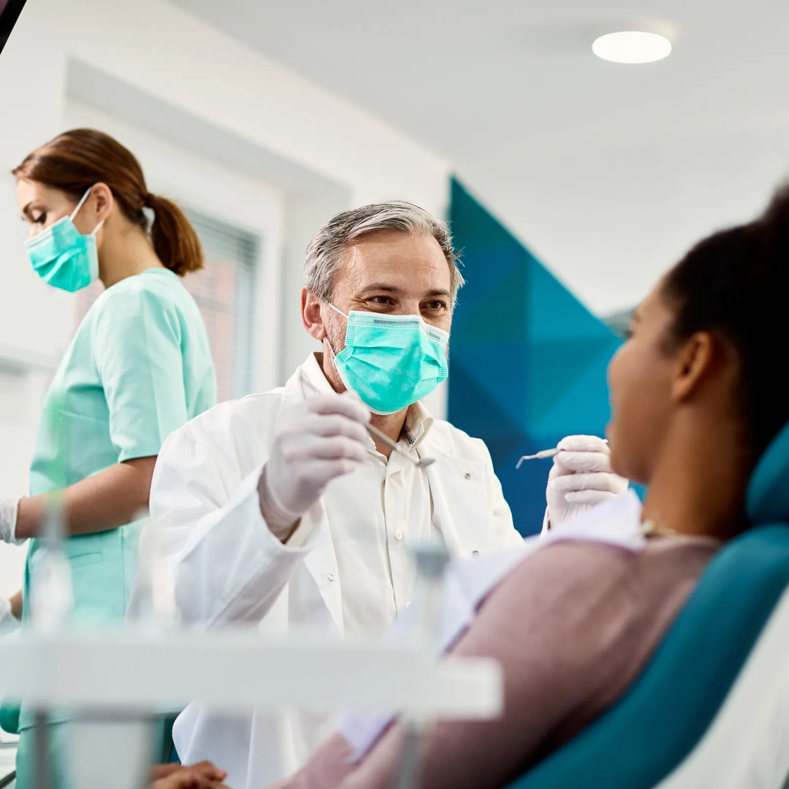 Dentist with patient in clinic, dental instruments and staff in background, promoting oral health and care.