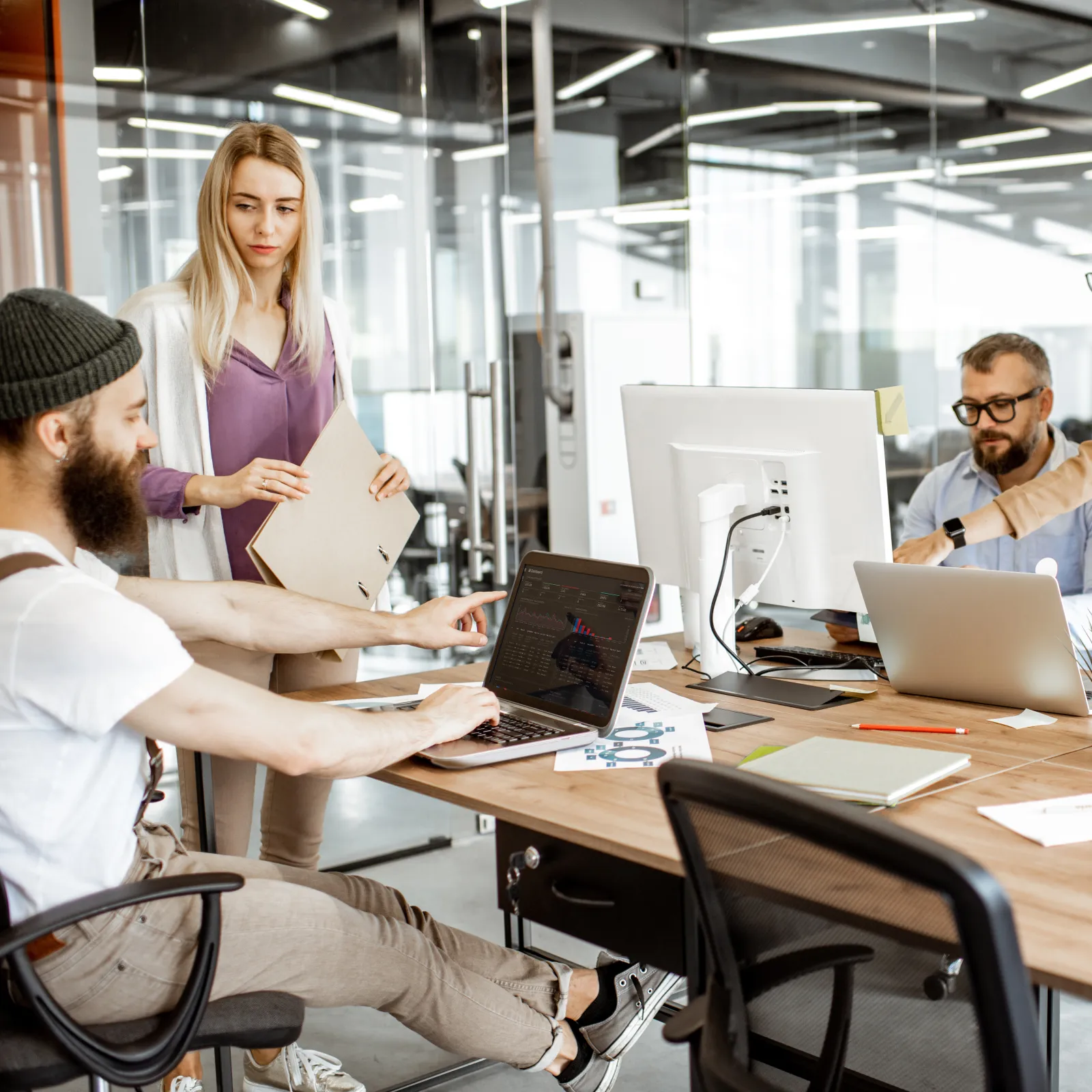 A team collaborates in a modern office, discussing projects at a wooden conference table with laptops.