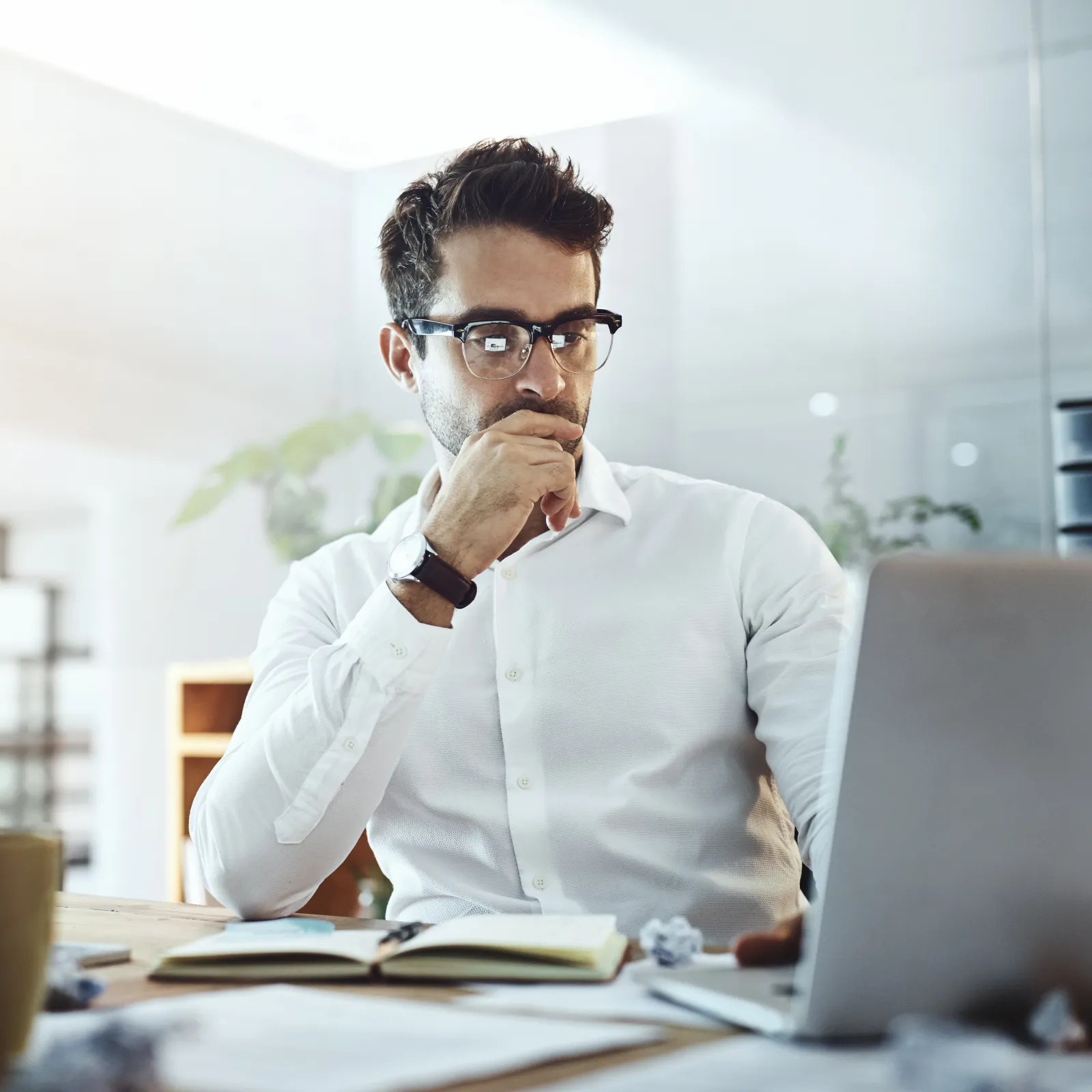 Thoughtful man in glasses working on laptop at a modern office desk.
