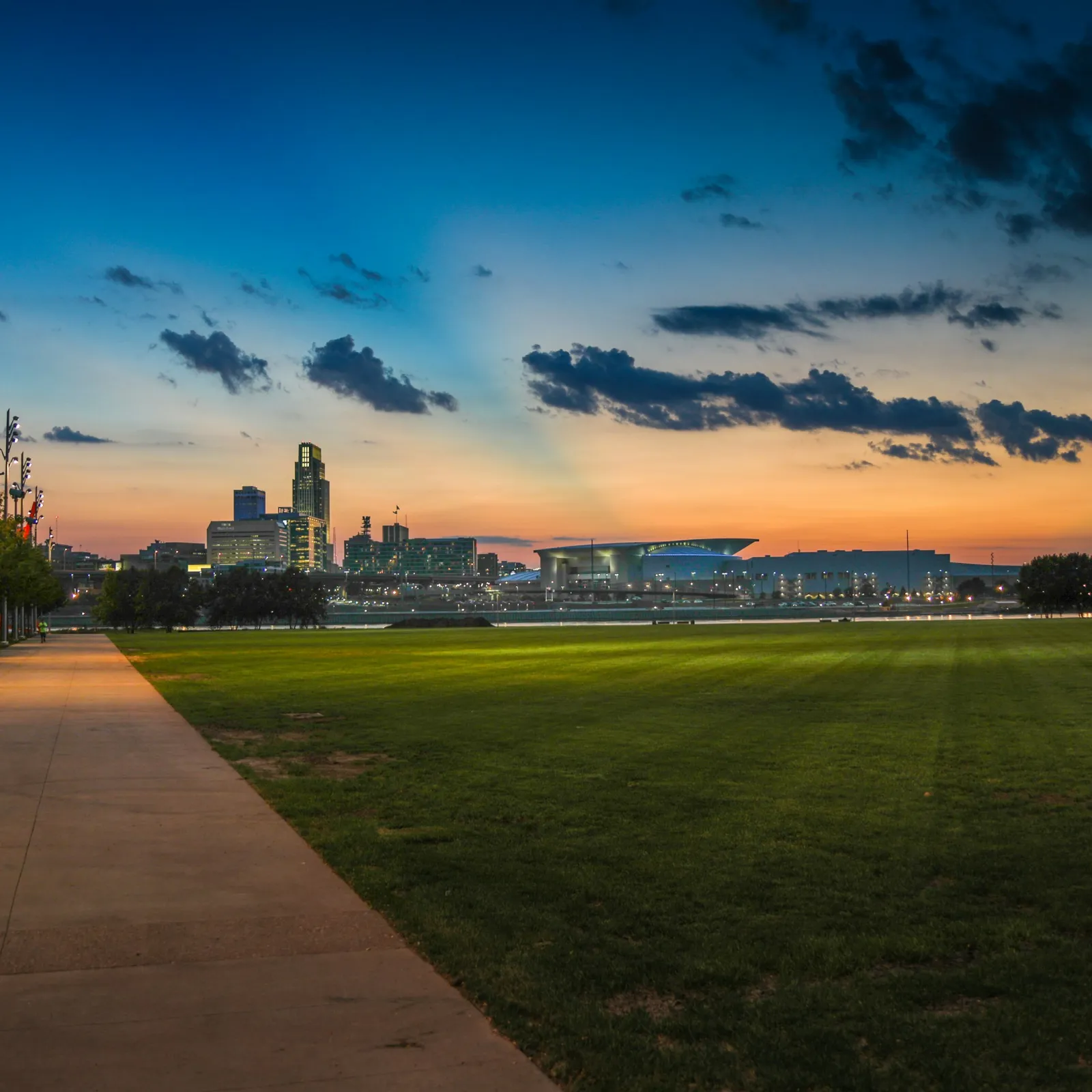 City skyline at sunset viewed from a park with walkway, lampposts, and green grass under a colorful sky.