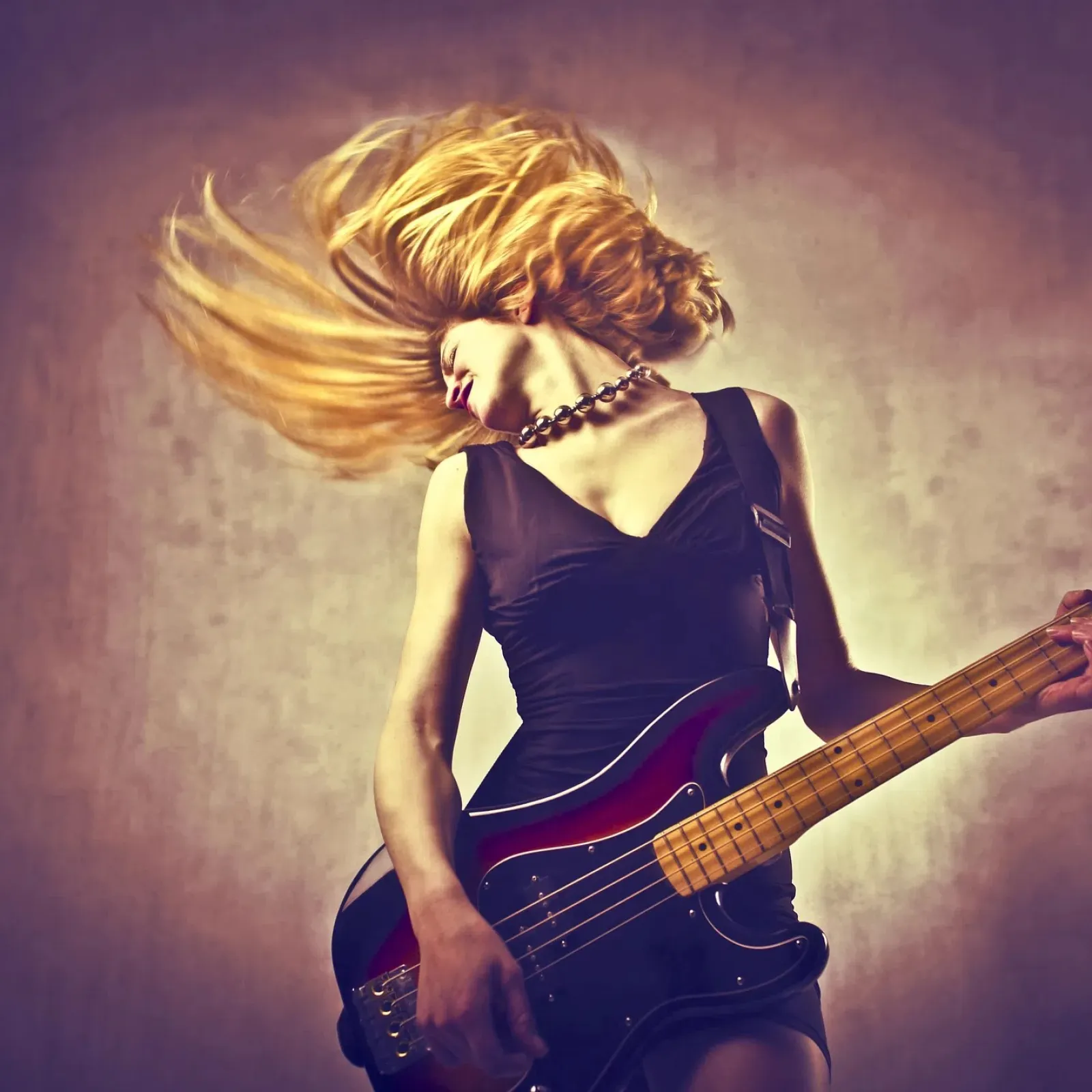 Blonde woman passionately playing electric bass guitar with hair flying against grungy background lighting.
