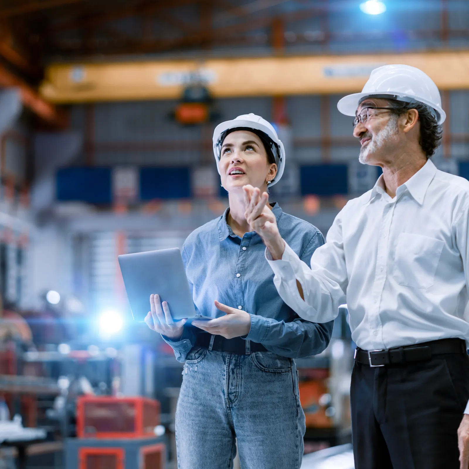 Two engineers in hard hats discussing plans inside an industrial factory with machinery in the background.